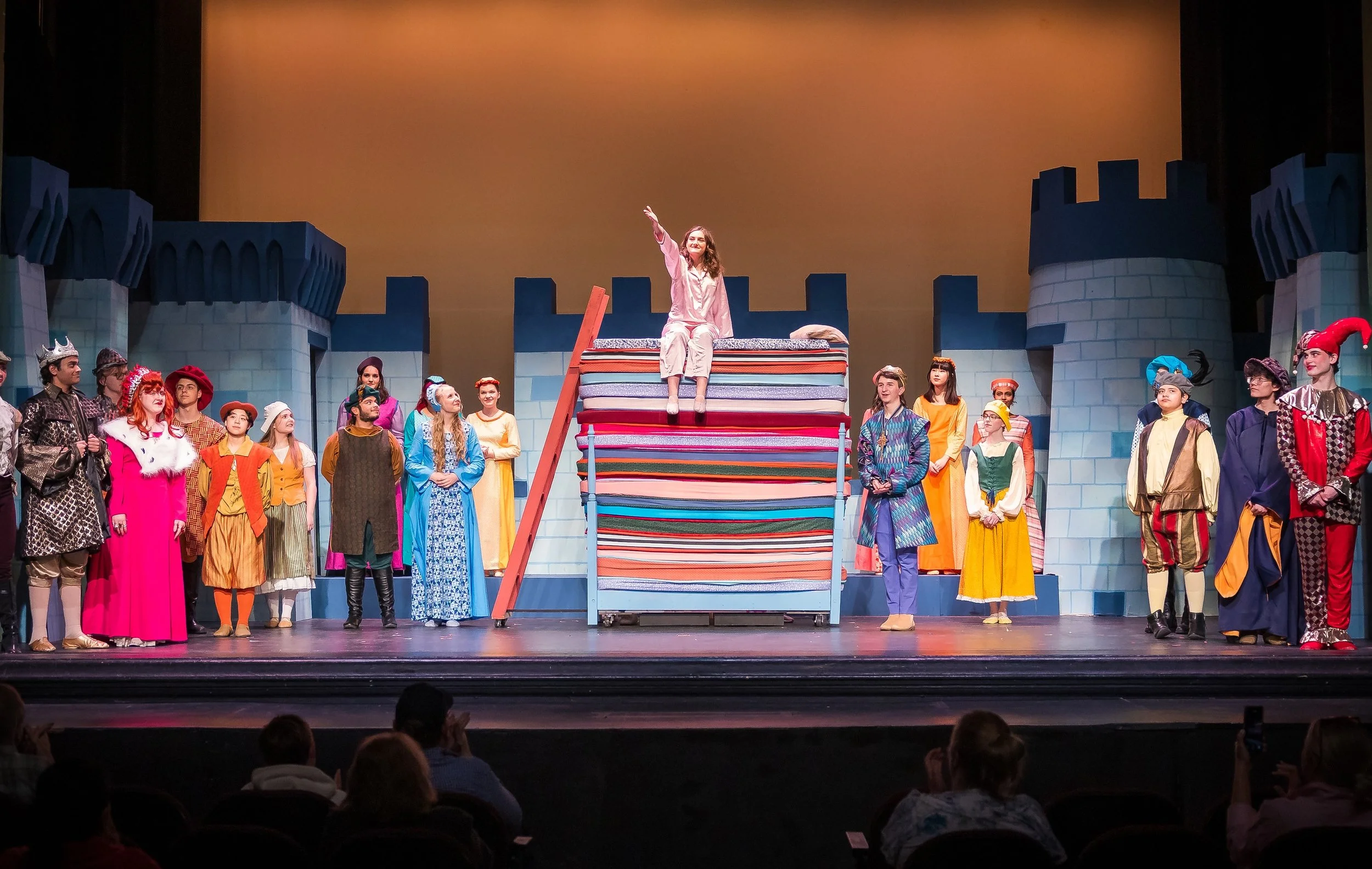 Actors on a stage in a theatrical production, with a castle backdrop, featuring a woman sitting on a pile of colorful blankets raising one arm, surrounded by other performers in medieval costumes.
