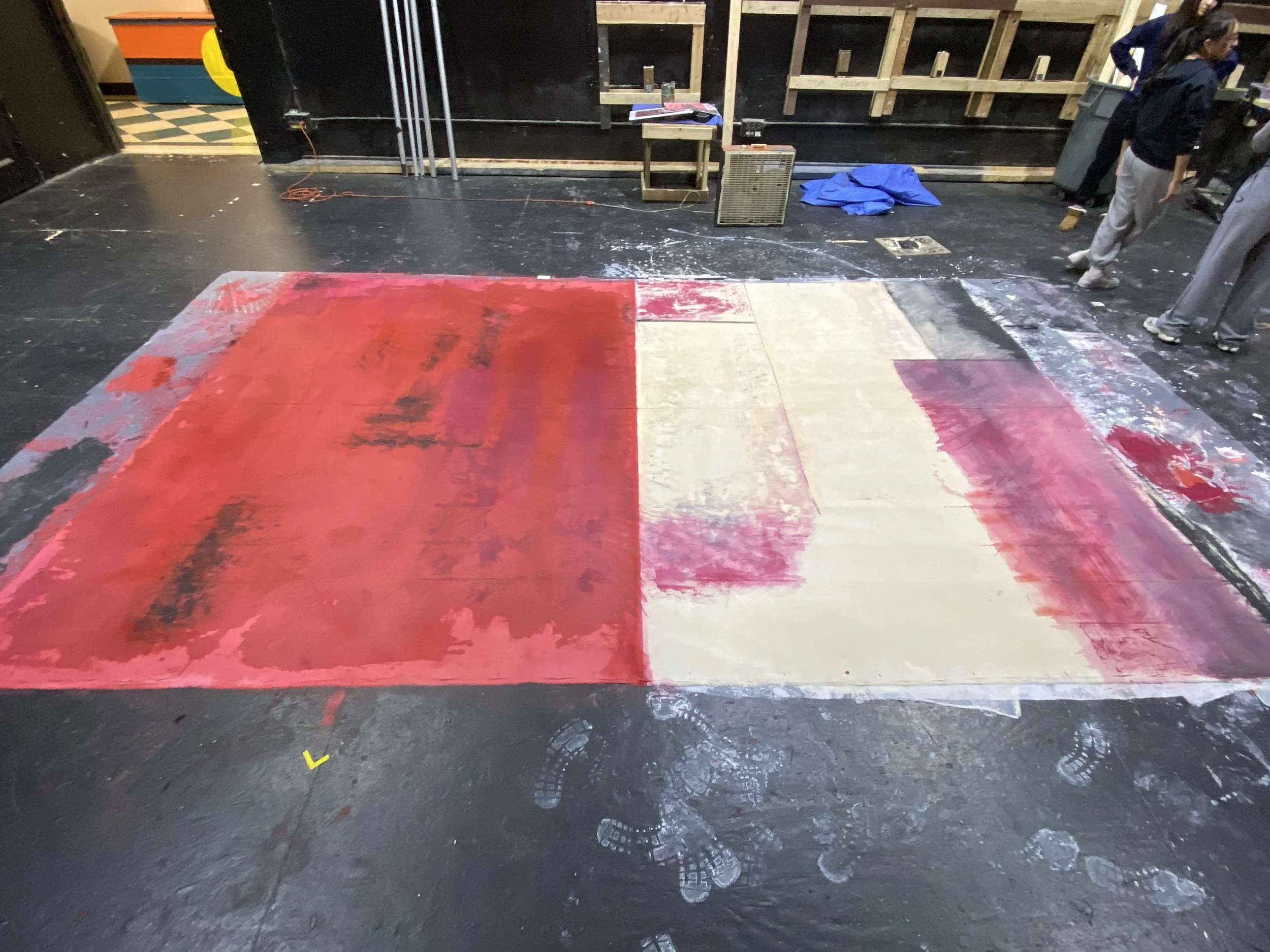 An indoor scene showing a floor with a partially painted red and beige backdrop. Several people are present at the right edge of the image, with some dusts and paint splatters on the floor.