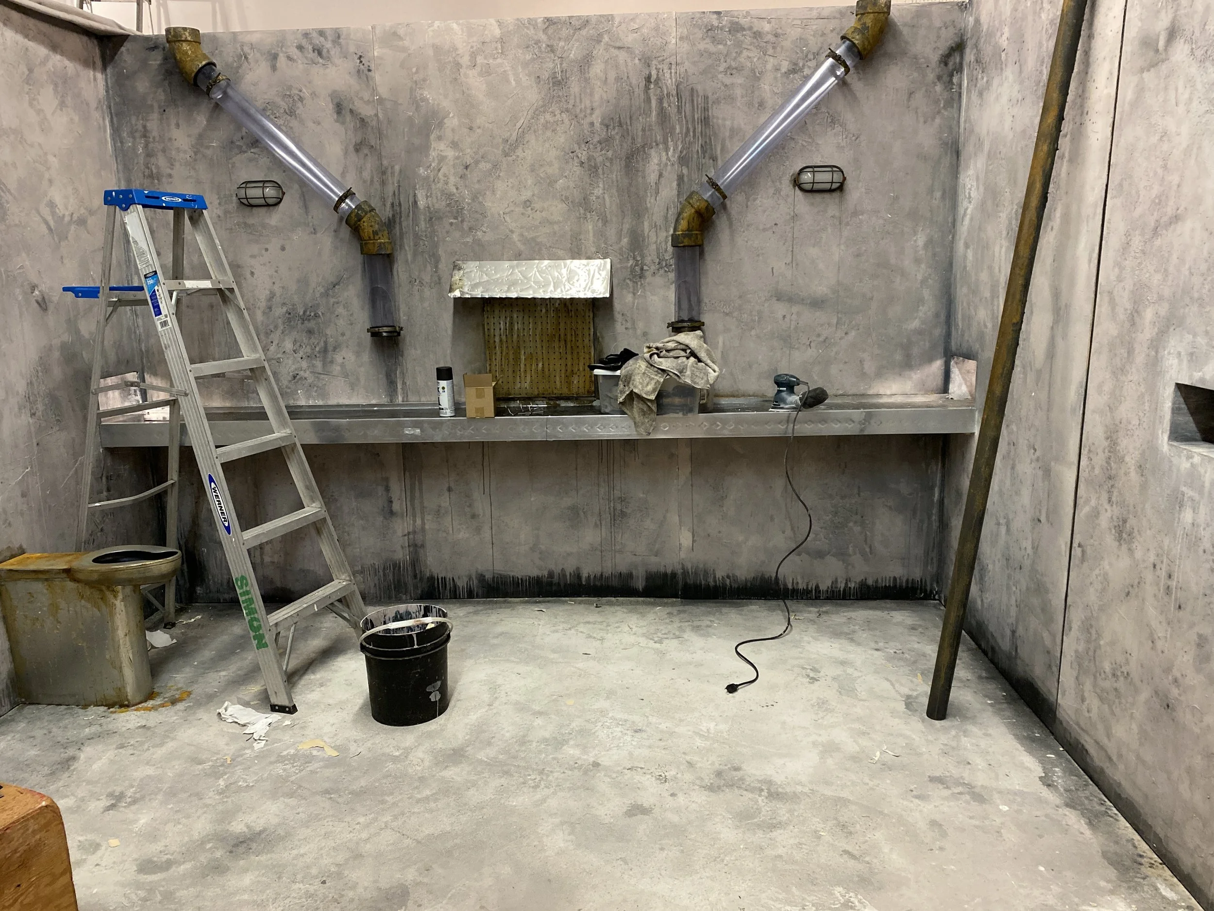Film set construction site with unfinished concrete walls and floor, yellow pipes and a metal work platform, ladder, trash can, and construction tools.