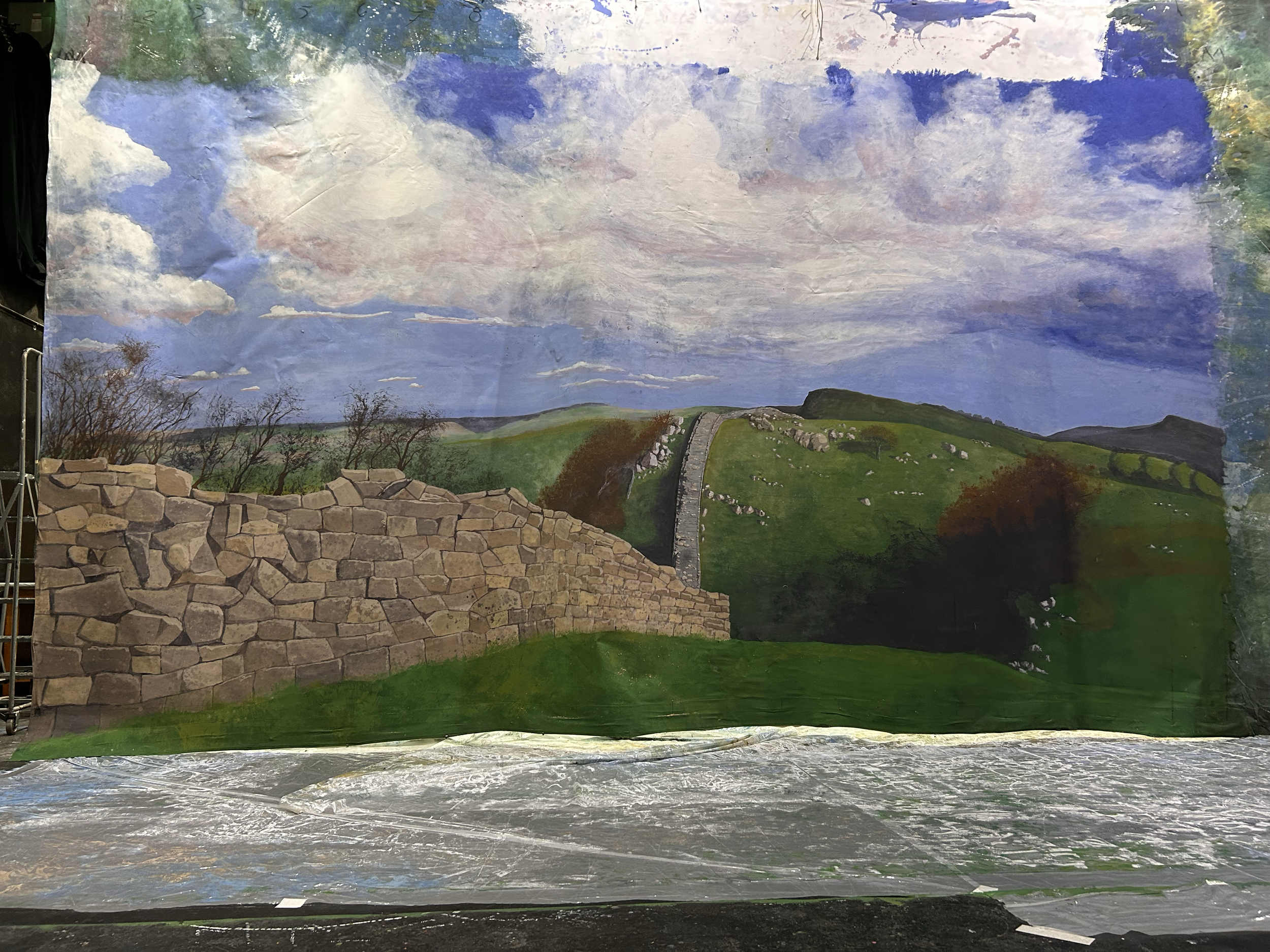 Theatrical backdrop painting of a landscape with a stone wall in the foreground, a hill with trees, and a sky filled with clouds.