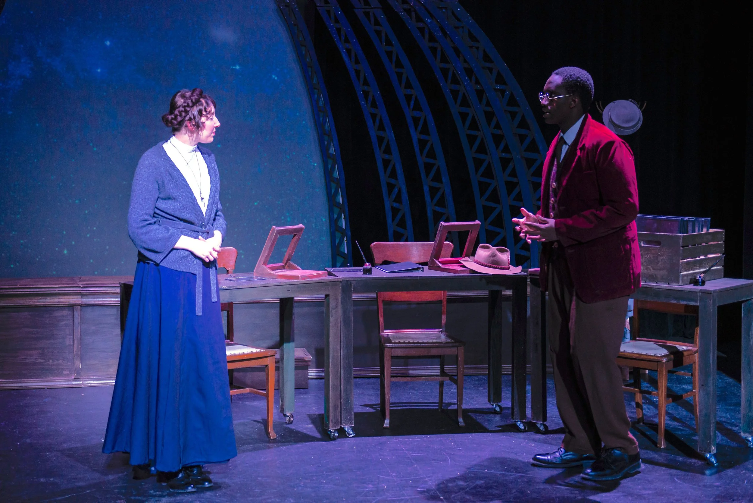 Two performers standing on a stage with a large blue starry backdrop, a table with various objects including a hat, and chairs in the background. The woman on the left has braided hair and is wearing a long skirt and cardigan, while the man on the ri