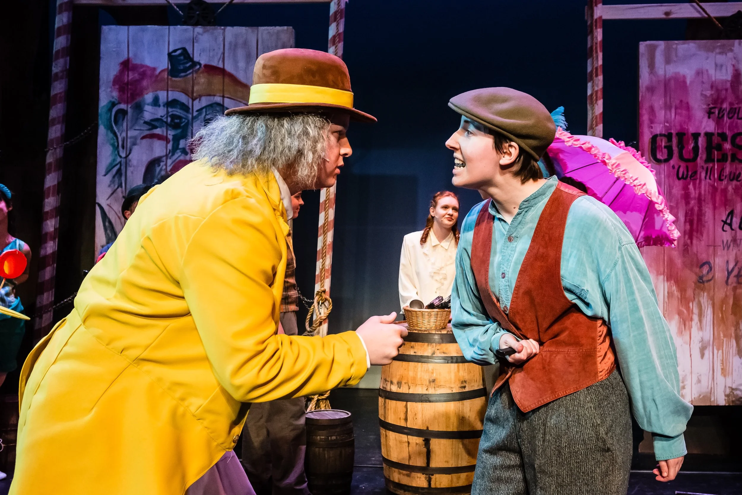 Two actors in costume face each other on stage, one wearing a yellow coat and brown hat, the other in a green shirt and brown vest, with a lady holding a pink parasol in the background.