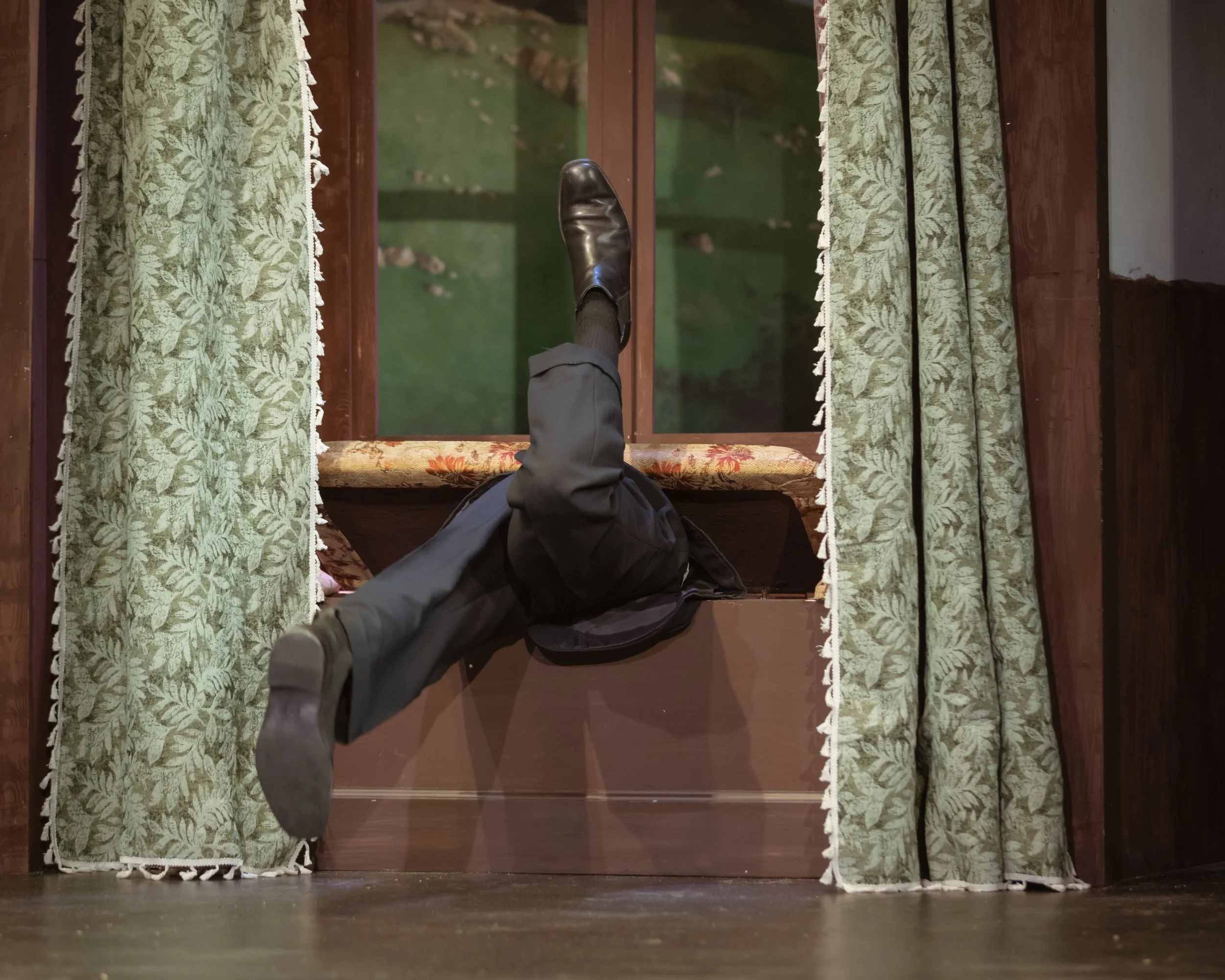 A theater set gag element showing a window seat with a secret trap door and green floral curtains on the sides. An actor is trapped under the window seat top, and their legs are flailing in the air.