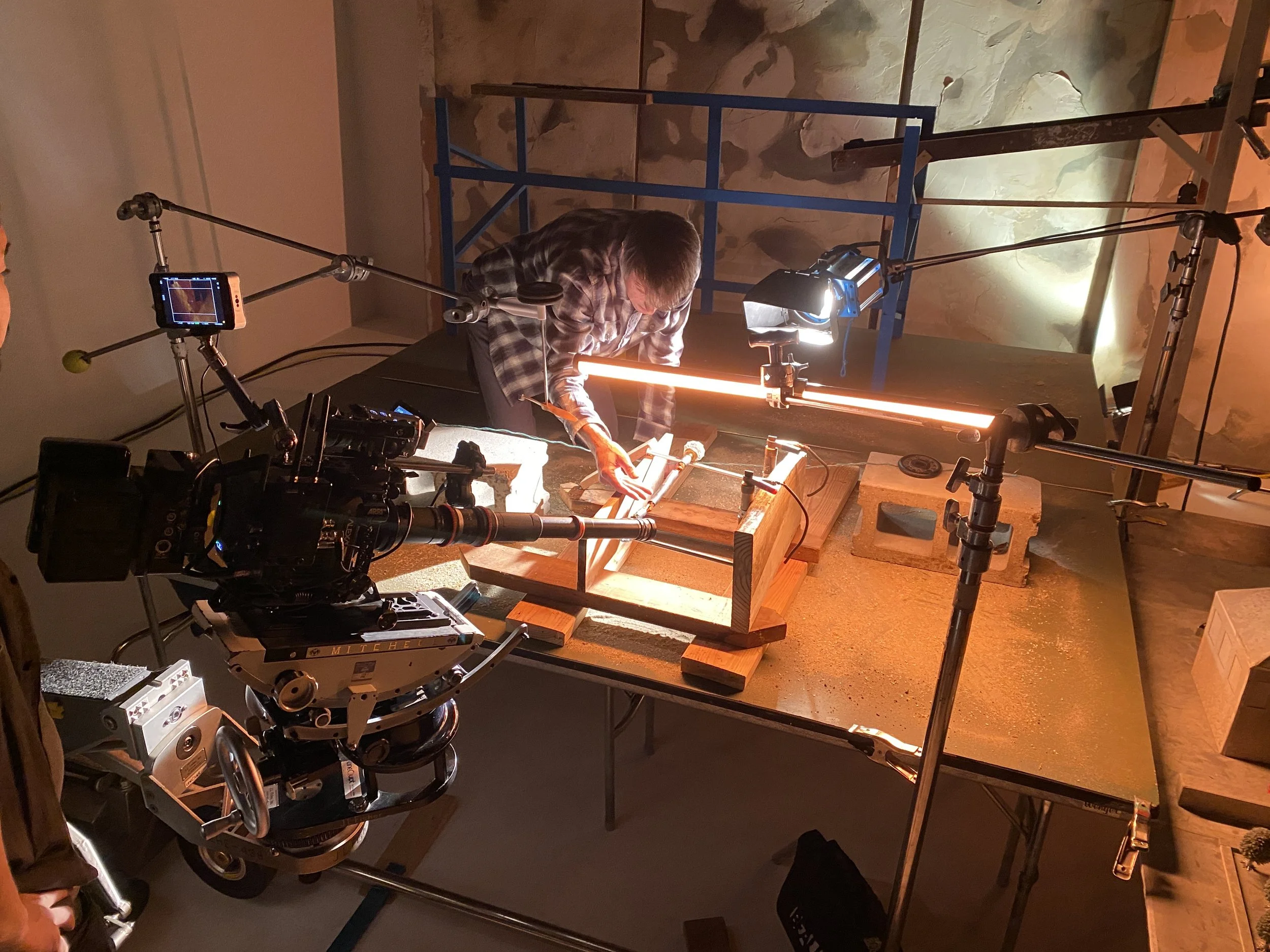 A person operating a camera on a tripod capturing footage of a person working with wooden materials under blue studio lighting. The scene is a movie filming setup in a room with textured, painted walls.