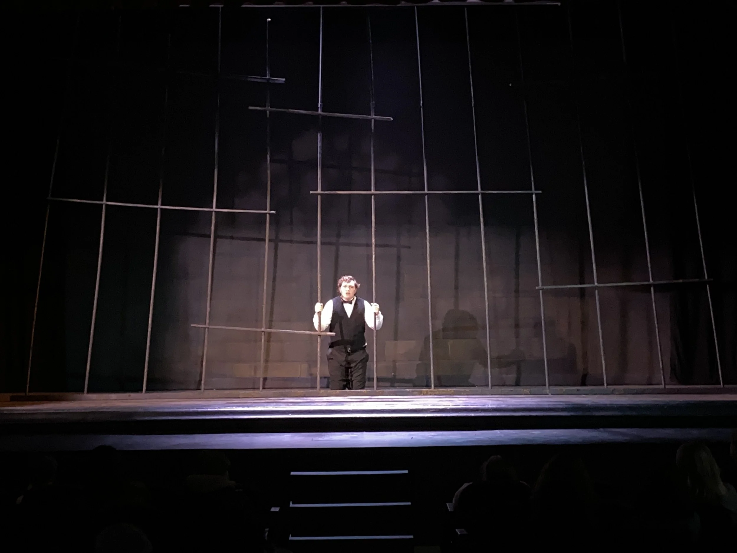 A performer on stage holding onto the bars of a stylized prison cell, with a dark background and grid-like structure behind them.