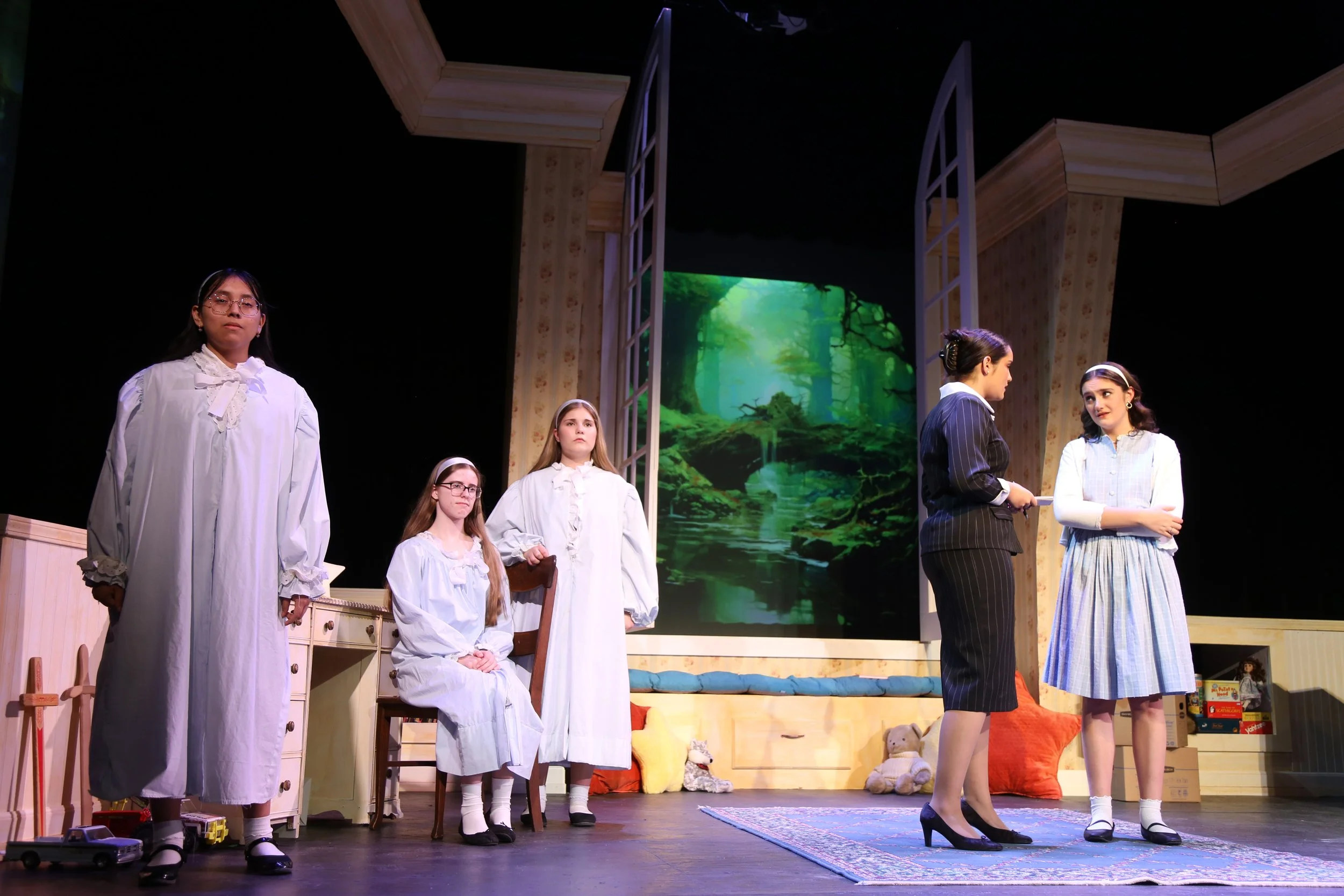 The image shows a stage scene from a play with six young women, some standing and some seated, in vintage or period costumes. The background features a scenic backdrop of a forest with lush green trees and a small waterfall. The stage is decorated wi