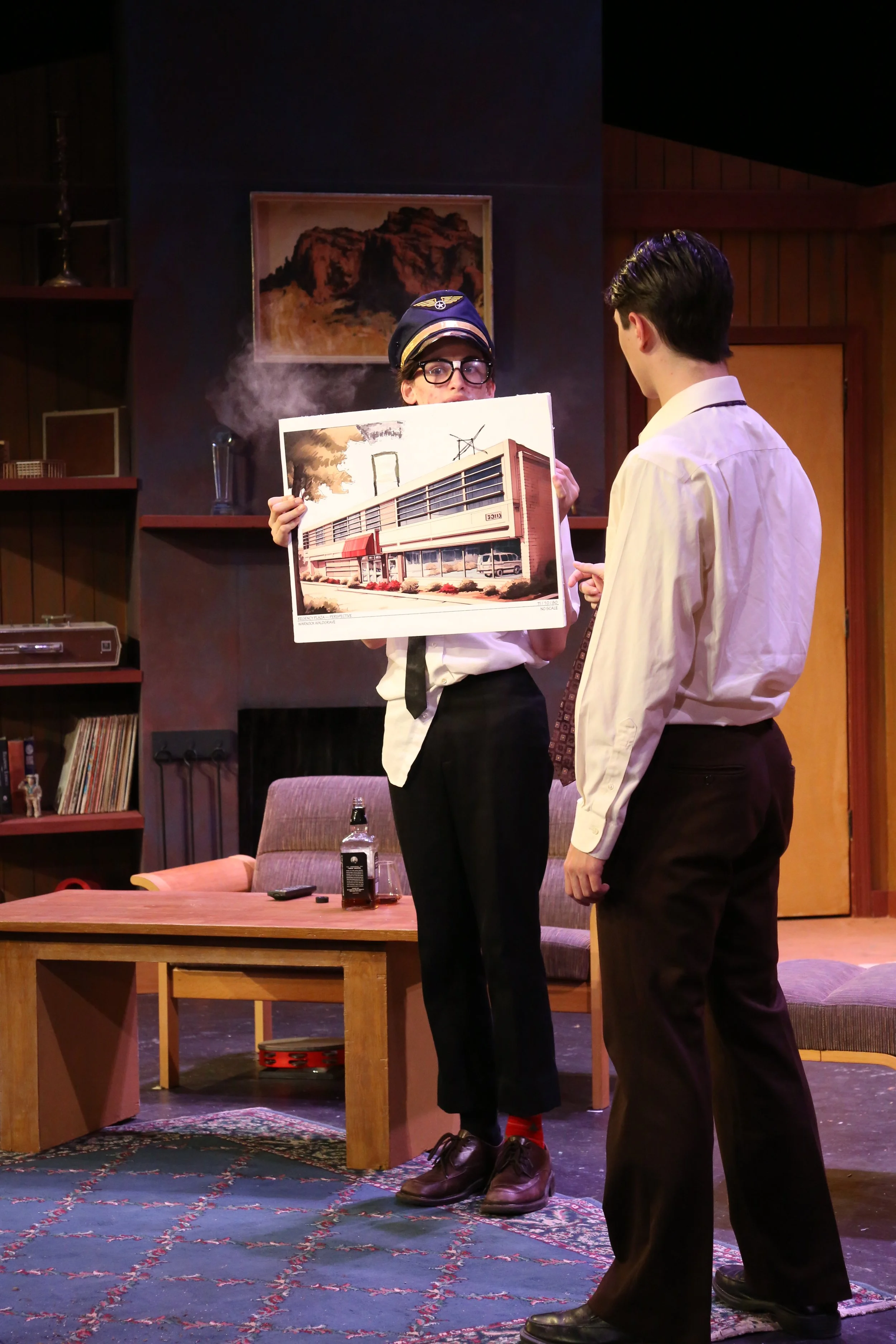 Two young men in a stage play are standing inside a room with wooden paneling, a bookshelf, and a painting of a mountain on the wall. One man, wearing a pilot's hat and glasses, is holding a large illustrated paper depicting a building and a tree, wh