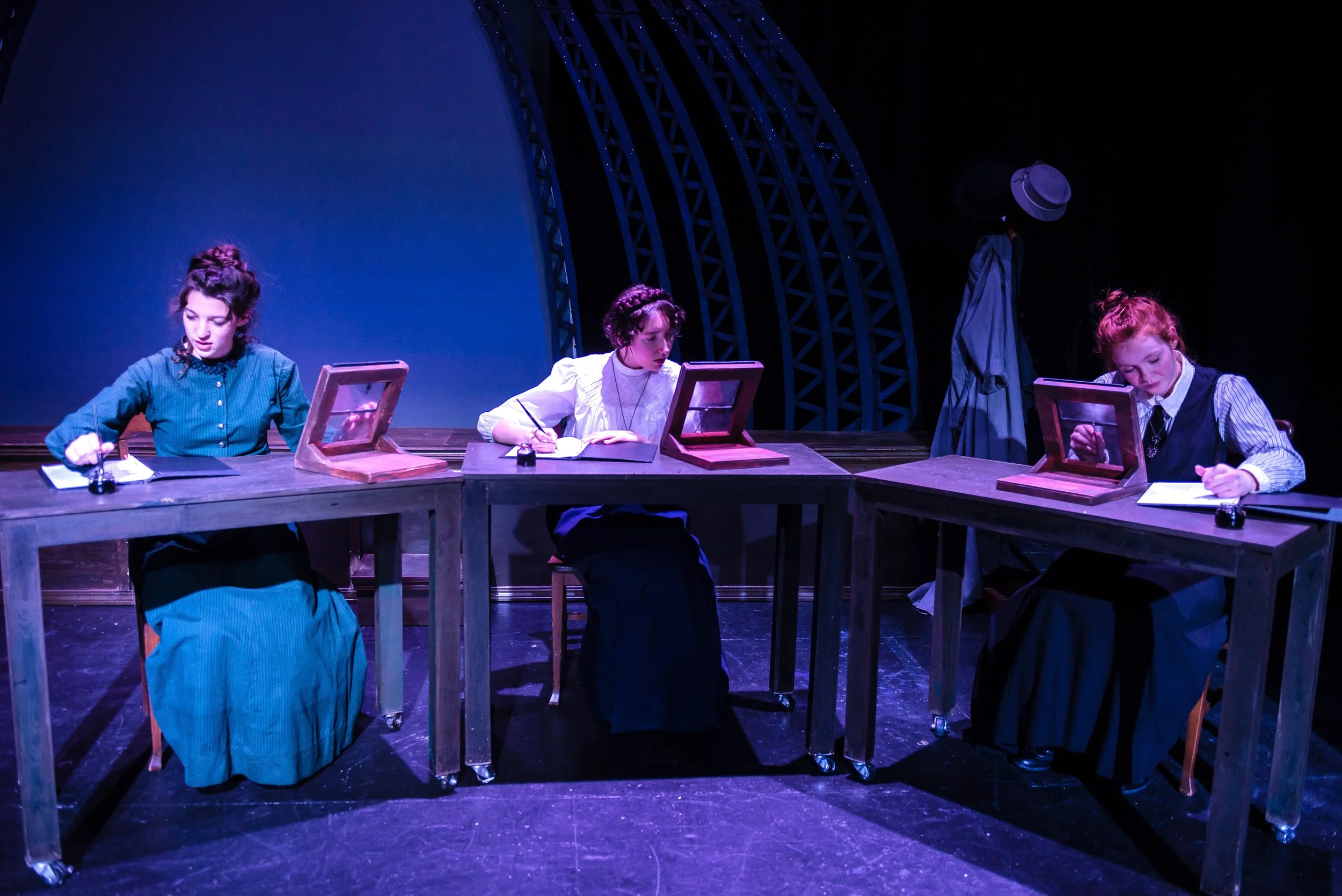 Three women in old-fashioned clothing sitting at a wooden table on stage, writing with ink pens, with small mirrors in front of them and a person in costume standing behind them, under stage lighting.