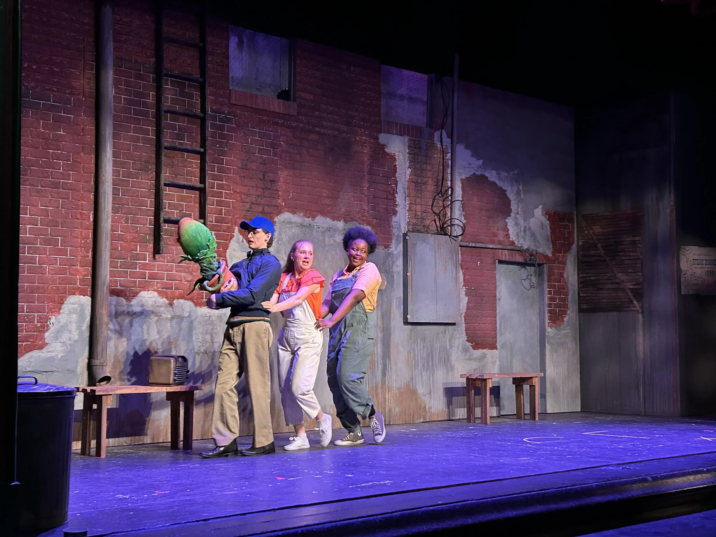 Four actors dancing on a stage with a brick wall backdrop. One actor holds a puppet plant, and all are dressed in casual clothing.