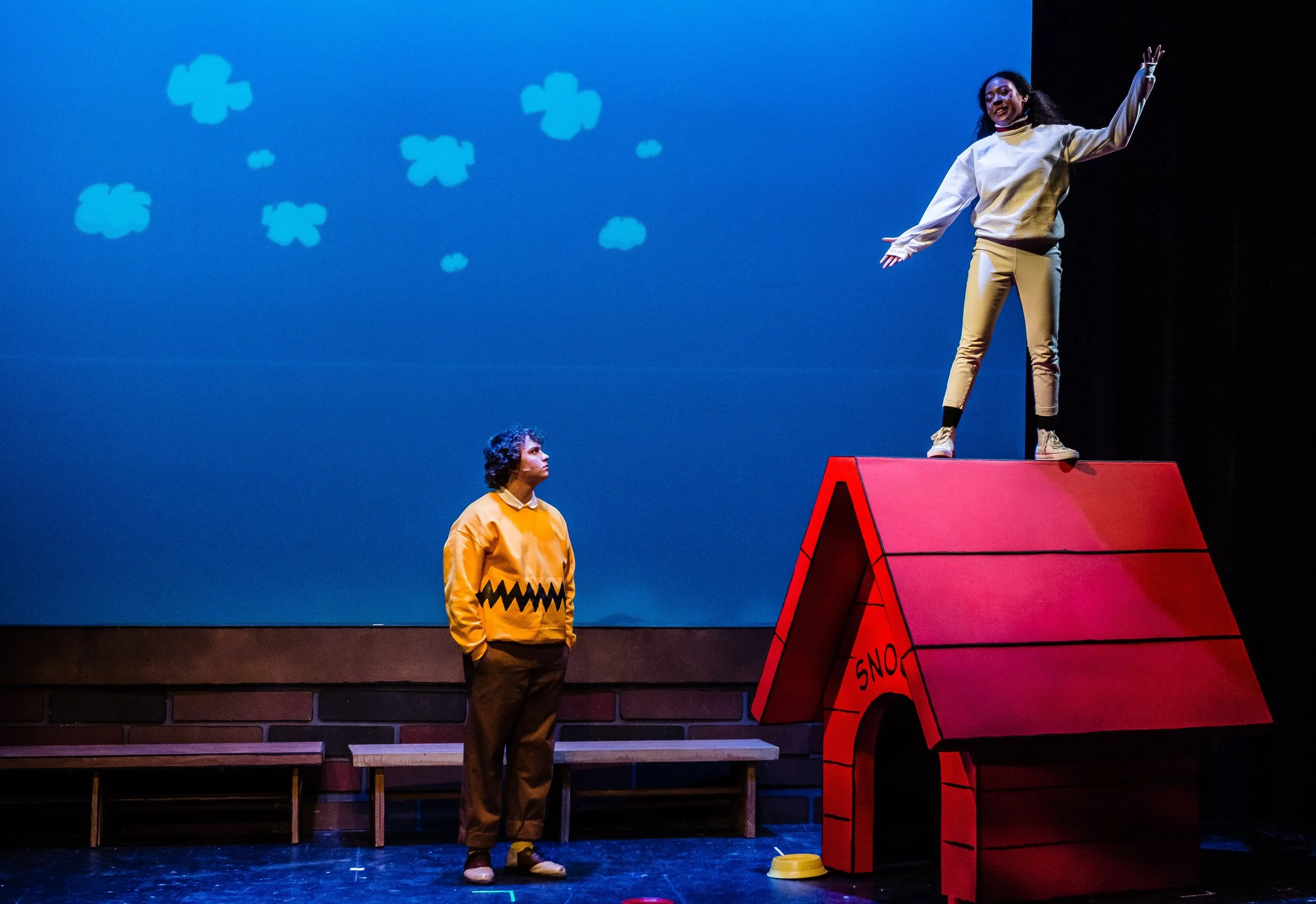 Two performers on a stage, one standing in front of a colorful doghouse and the other on top of it, with a blue background featuring cloud shapes.