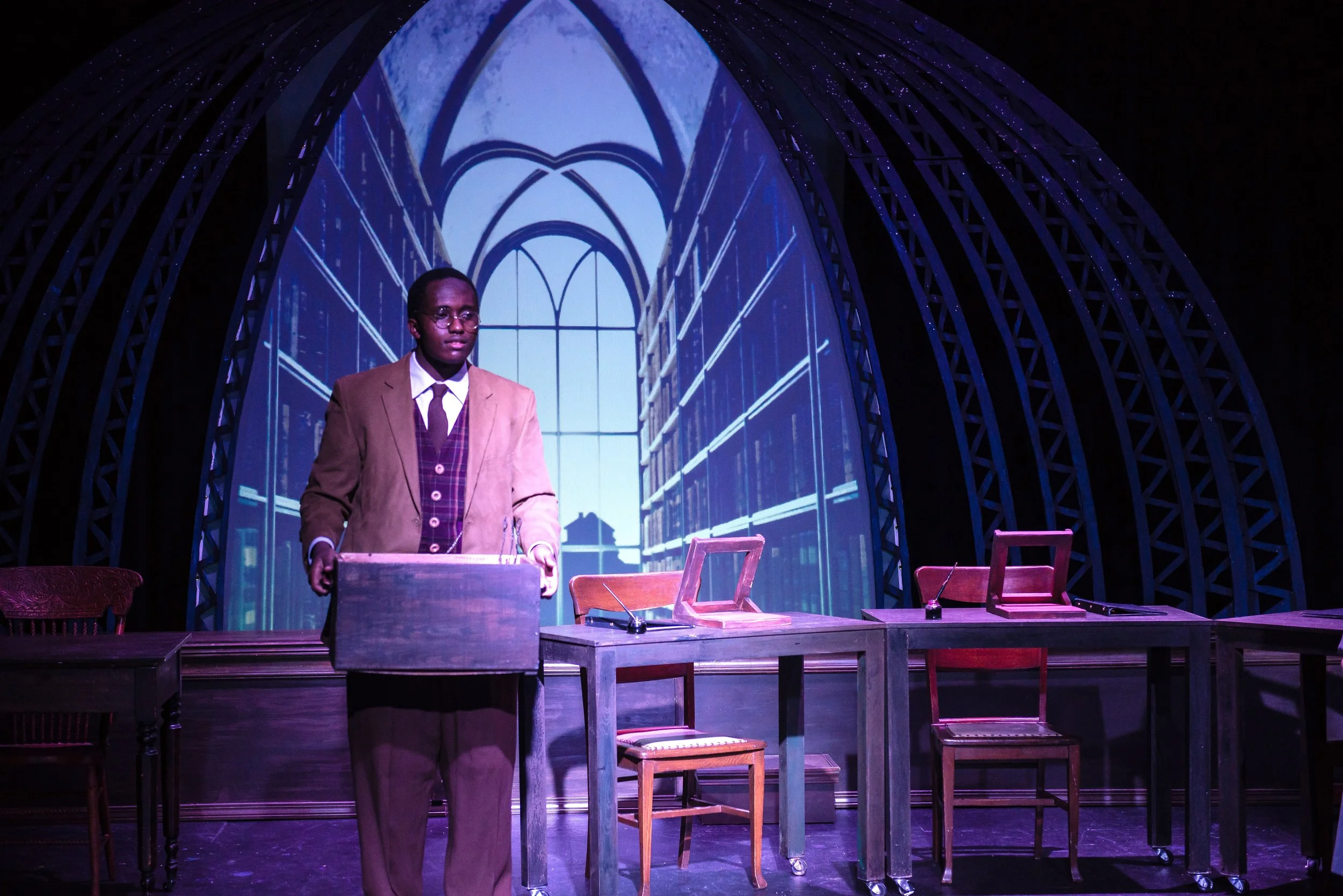 A man in a tan suit and glasses standing behind a wooden desk on a stage with chairs and a background of a large window and blue lighting.