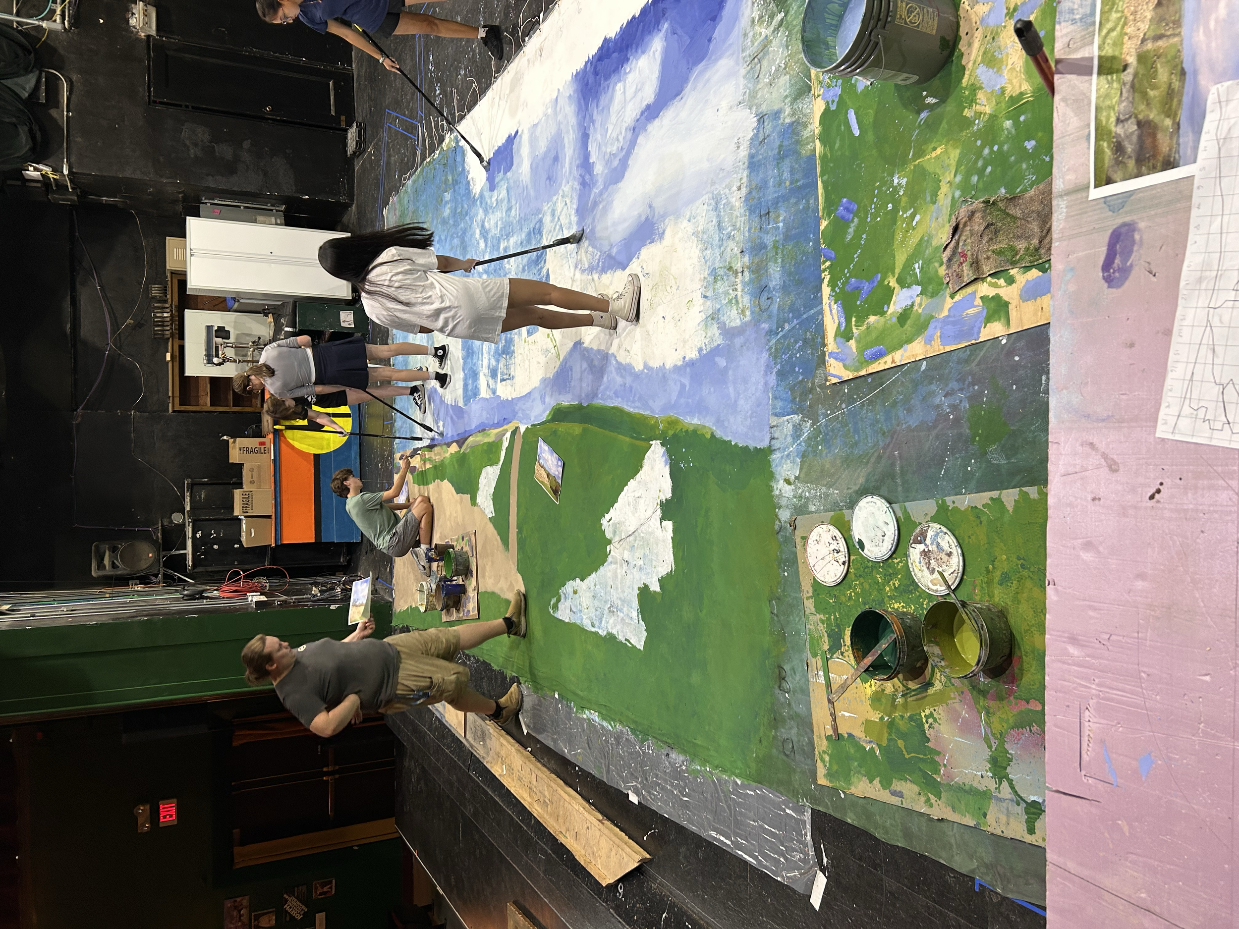 Several people working together on a large landscape painting with mountains and fields, using brushes and paints in an indoor studio.