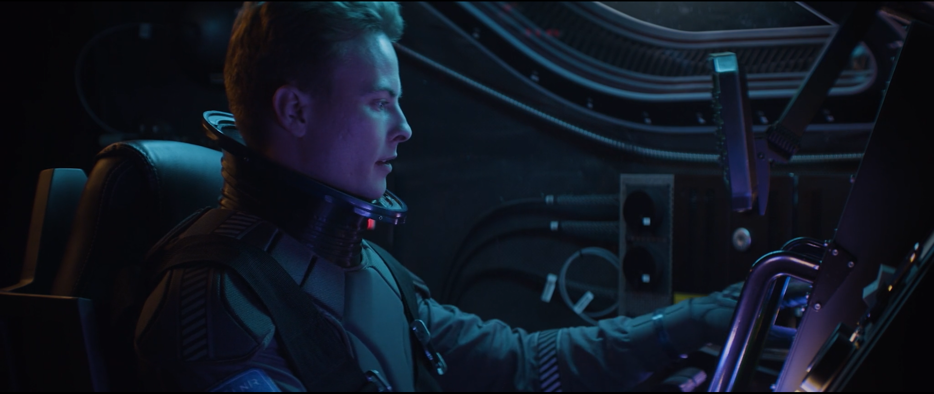 A person in a space suit sitting at a control panel in a spacecraft, facing a screen.