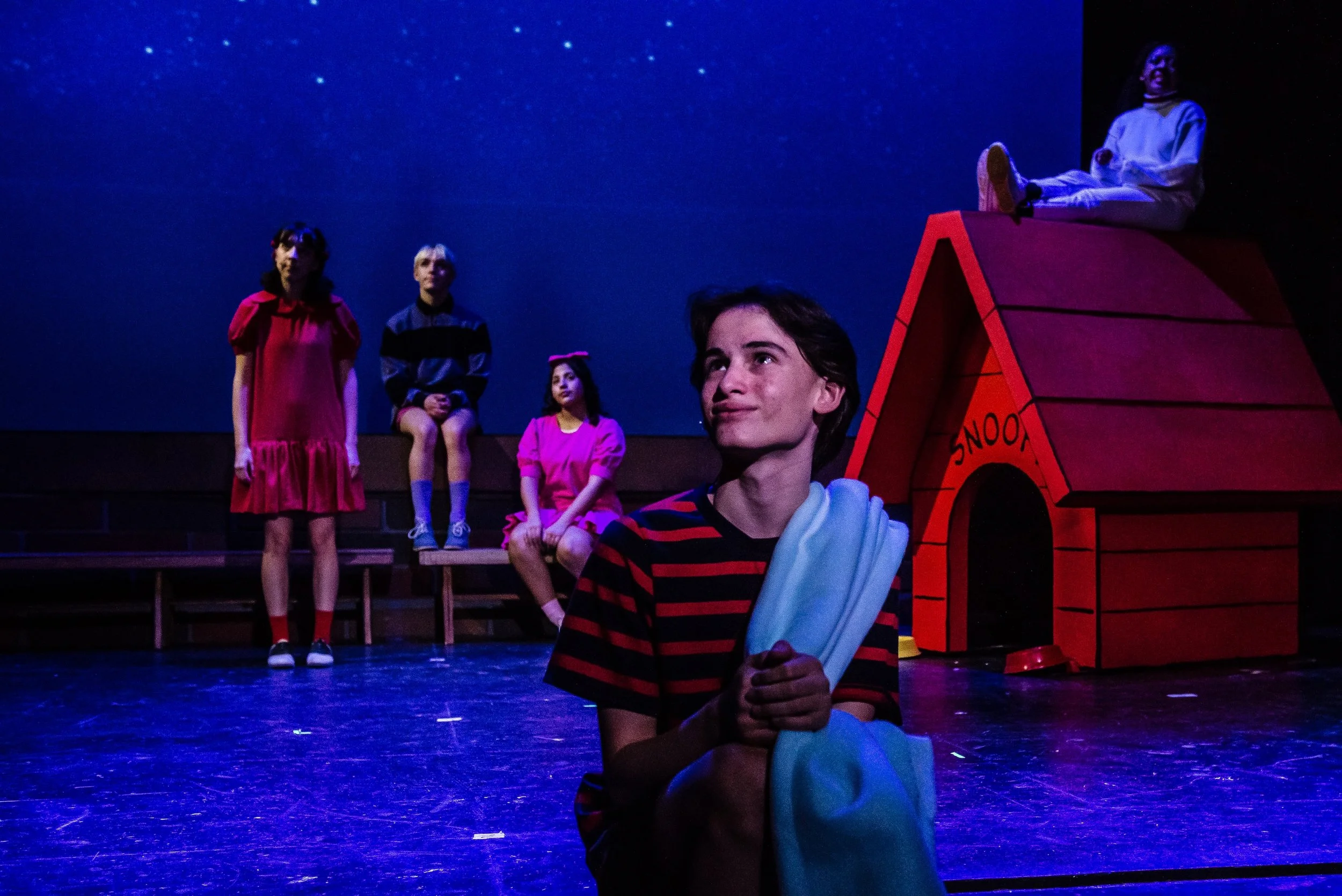 A young actor with dark hair is sitting on the stage, holding a blue child's blanket, surrounded by children in colorful costumes, with a starry background and a red doghouse labeled 'SNOOPY'.