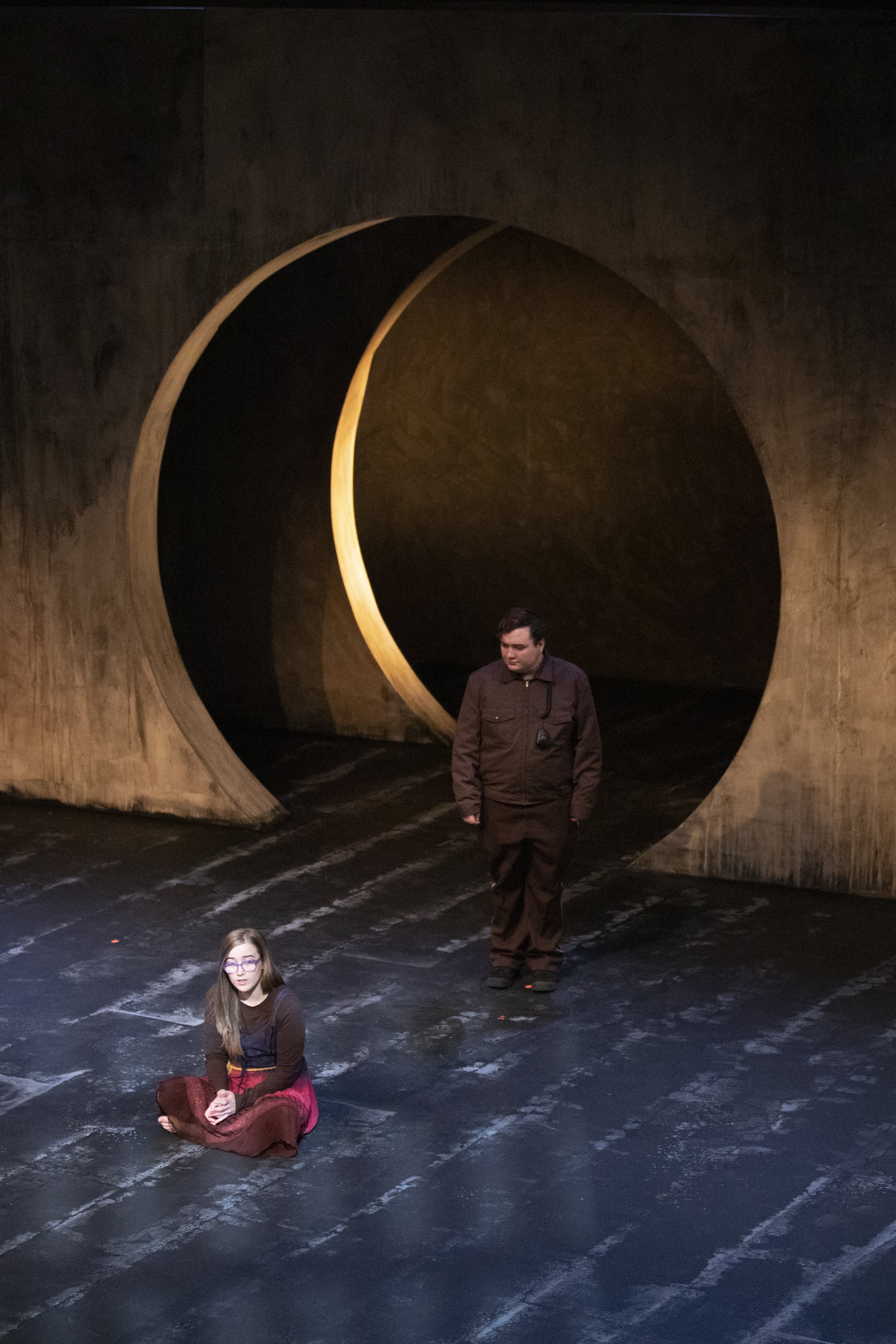 A stage with a large, circular, tunnel-like structure with a golden light inside. Two people are on the stage: a woman sitting cross-legged in the foreground, and a man walking behind her, dressed in dark clothing. The stage floor has black and white