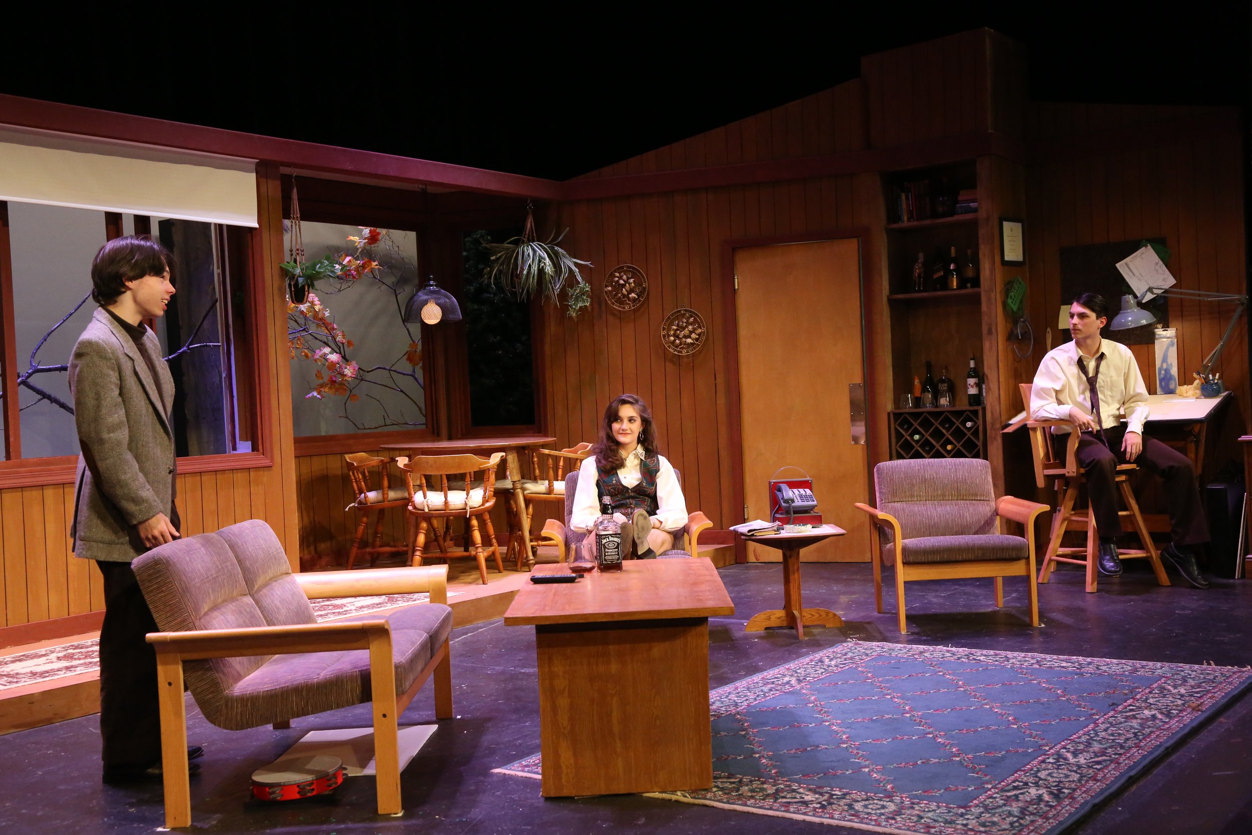 The image shows a stage set resembling a cozy living room with wooden walls. There are three people: one man standing on the left, a woman sitting in a wooden armchair, and a man sitting on a desk on the right. The room has various decorations, inclu