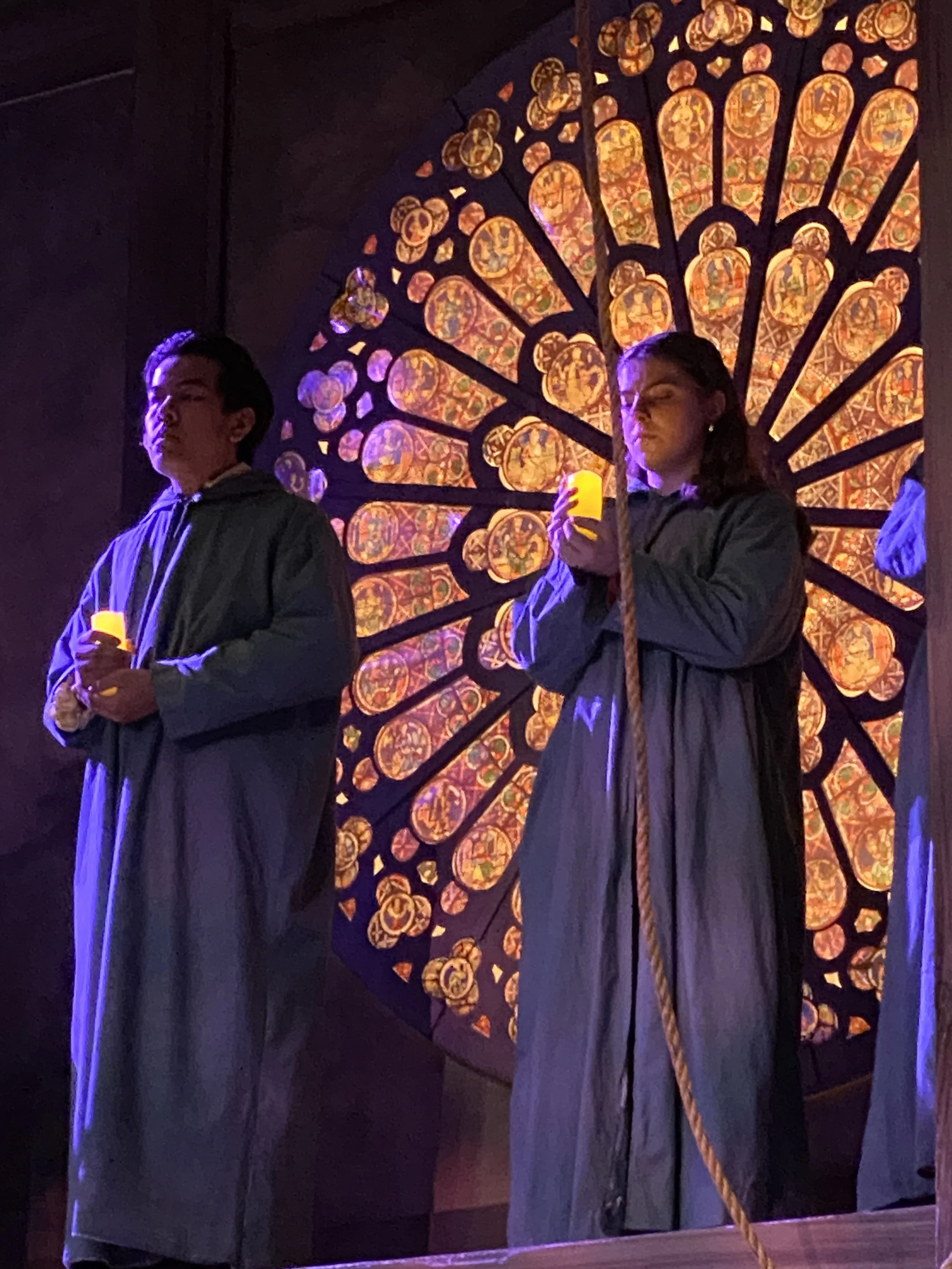 Two actors with dark hair wearing dark robes are holding yellow candles and standing in front of a large, illuminated stained glass window that features intricate, colorful circular patterns. The scene is dimly lit with a purple hue.