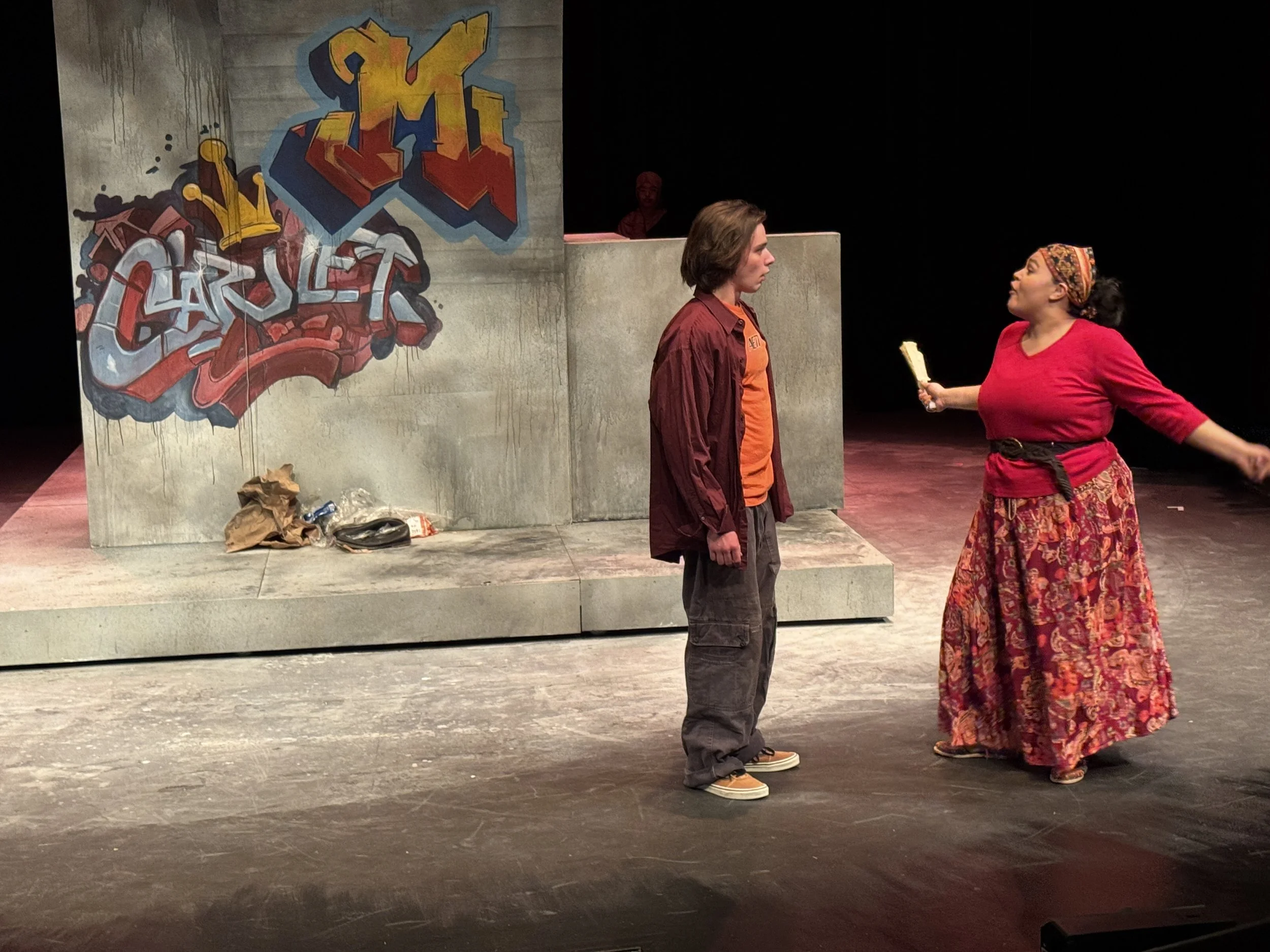 Two actors on stage. The actor on the left has shoulder-length hair and wears a maroon jacket with gray pants, looking serious. The actor on the right wears a vibrant red top, a colorful patterned skirt, and a headscarf, and speaking passionately. Be