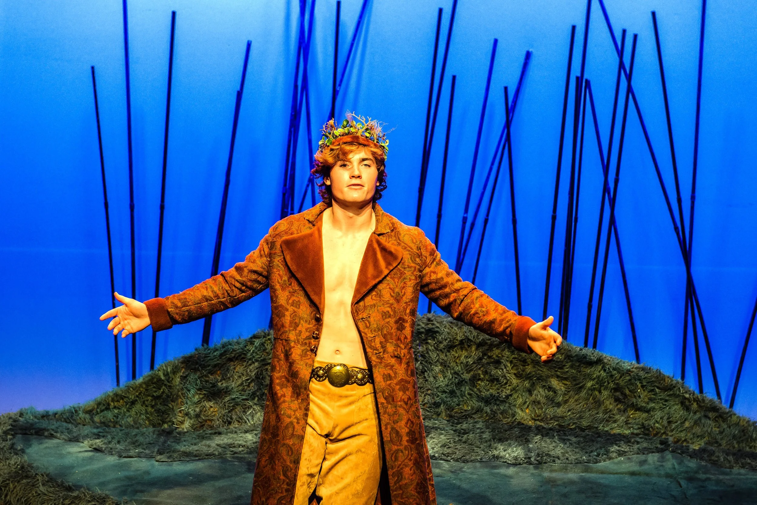 An actor playing Oberon with brown, curly hair wearing a floral crown and a patterned brown coat, standing with arms out to the sides against a blue background with black vertical lines and a textured foreground.