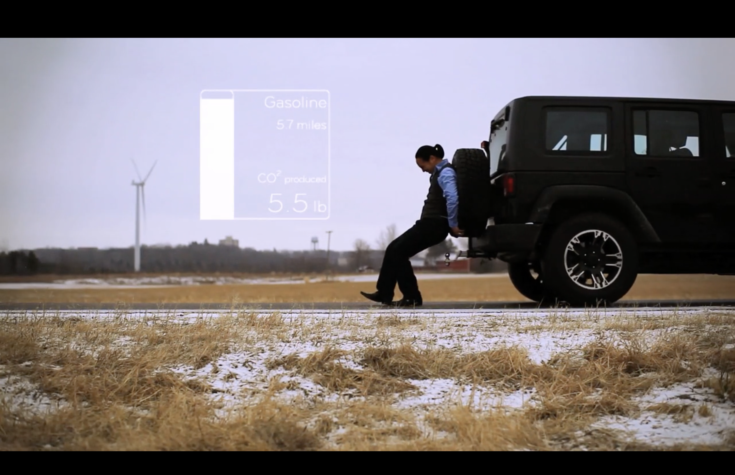 An actor in business attire is pushing a black SUV down a road with patches of snow. In the background, there is a wind turbine and a large infographic overlay showing statements about gasoline, miles, and CO2 emissions.