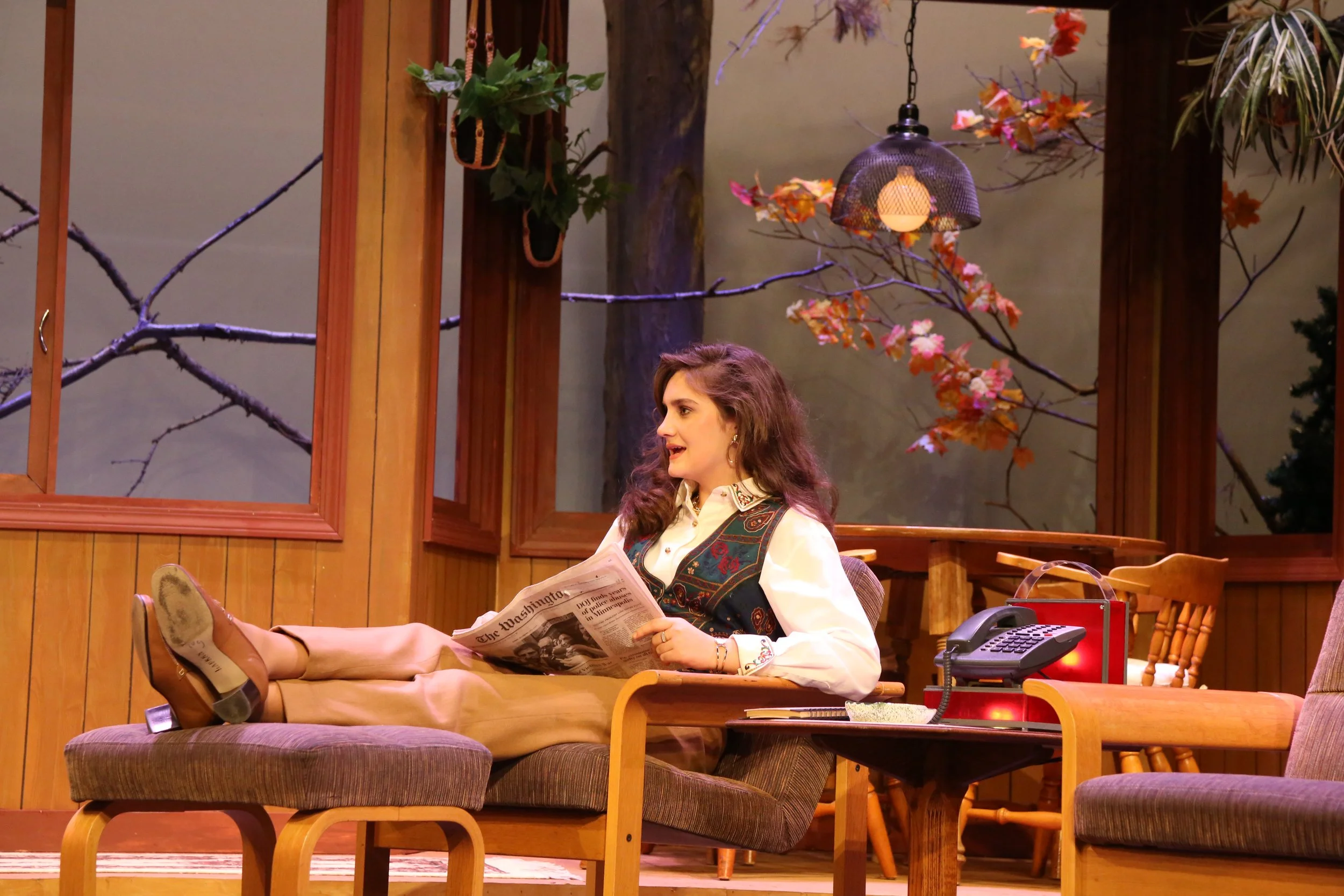 A woman with curly hair and earrings is sitting on a bench, reading a newspaper, while wearing beige shoes, in a cozy room with wood-paneled walls and large windows. The room has autumn-themed decorations, including branches with orange leaves, potte