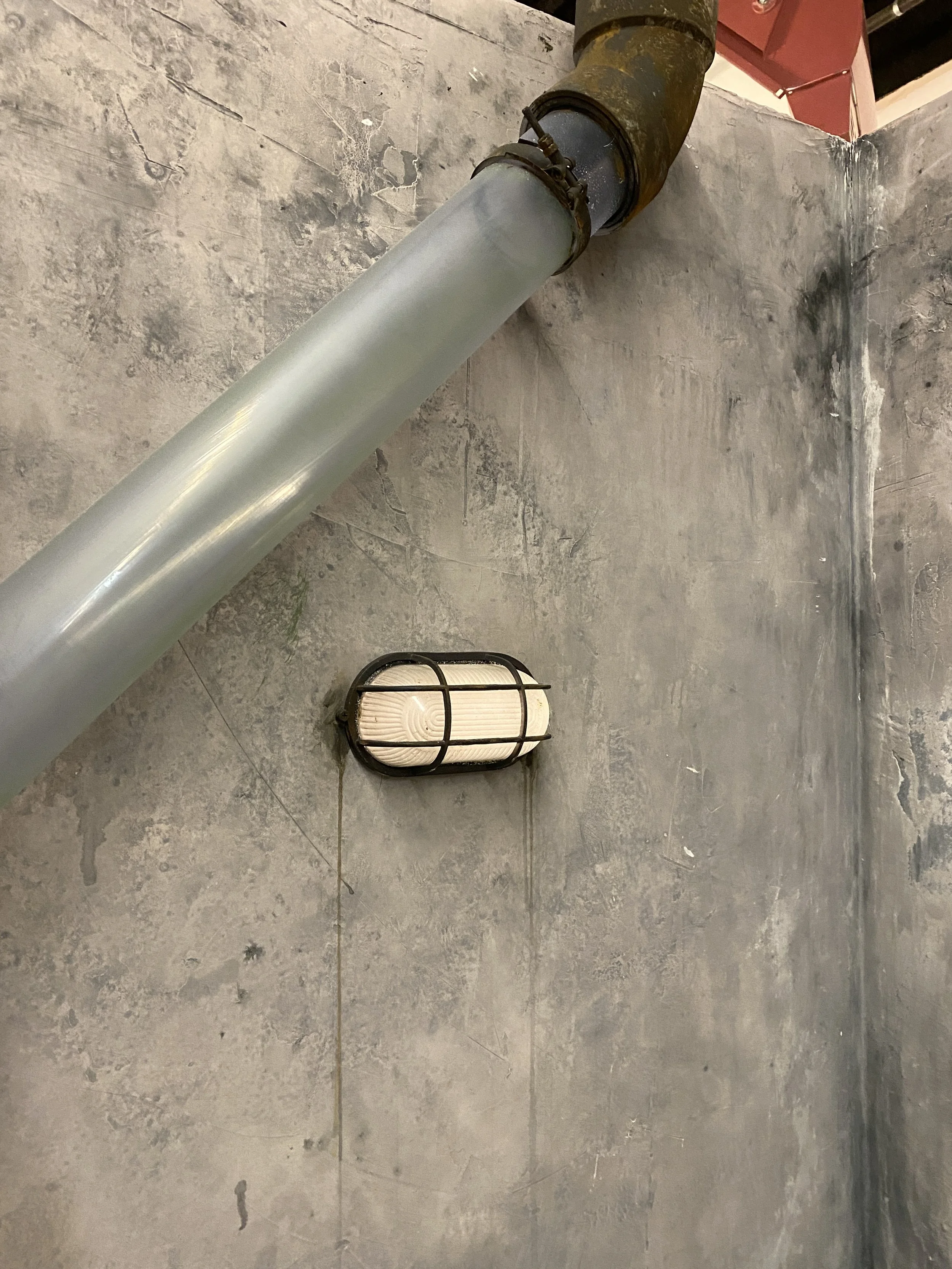 A large industrial pipe connected to a wall, with a filter or light fixture mounted nearby on the floor.