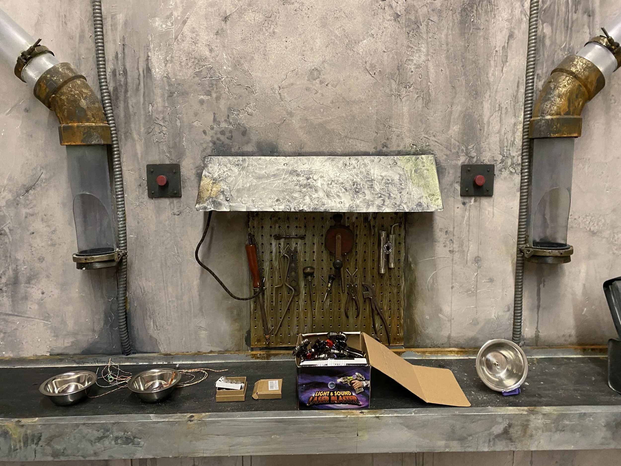 A film set with tools hanging on a pegboard, wires, metal bowls, and a box labeled "Light & Sound Laser Blaster," with pipes running along the wall.
