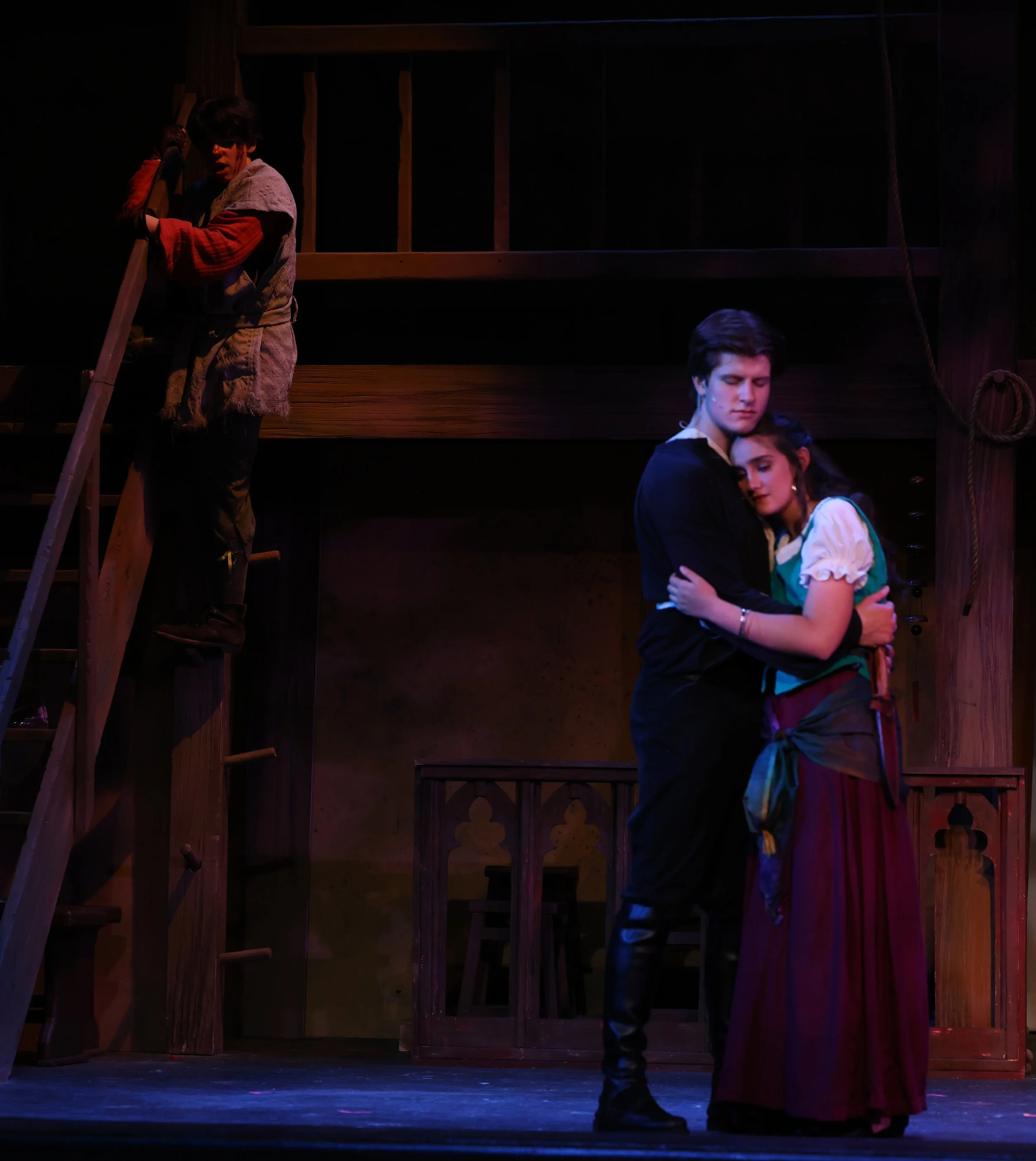 A theatrical scene with three actors on stage. One actor is ascending a wooden ladder, wearing a patchwork costume and fingerless gloves, looking surprised. Two actors are hugging in the foreground, one with a solemn expression and the other appearin