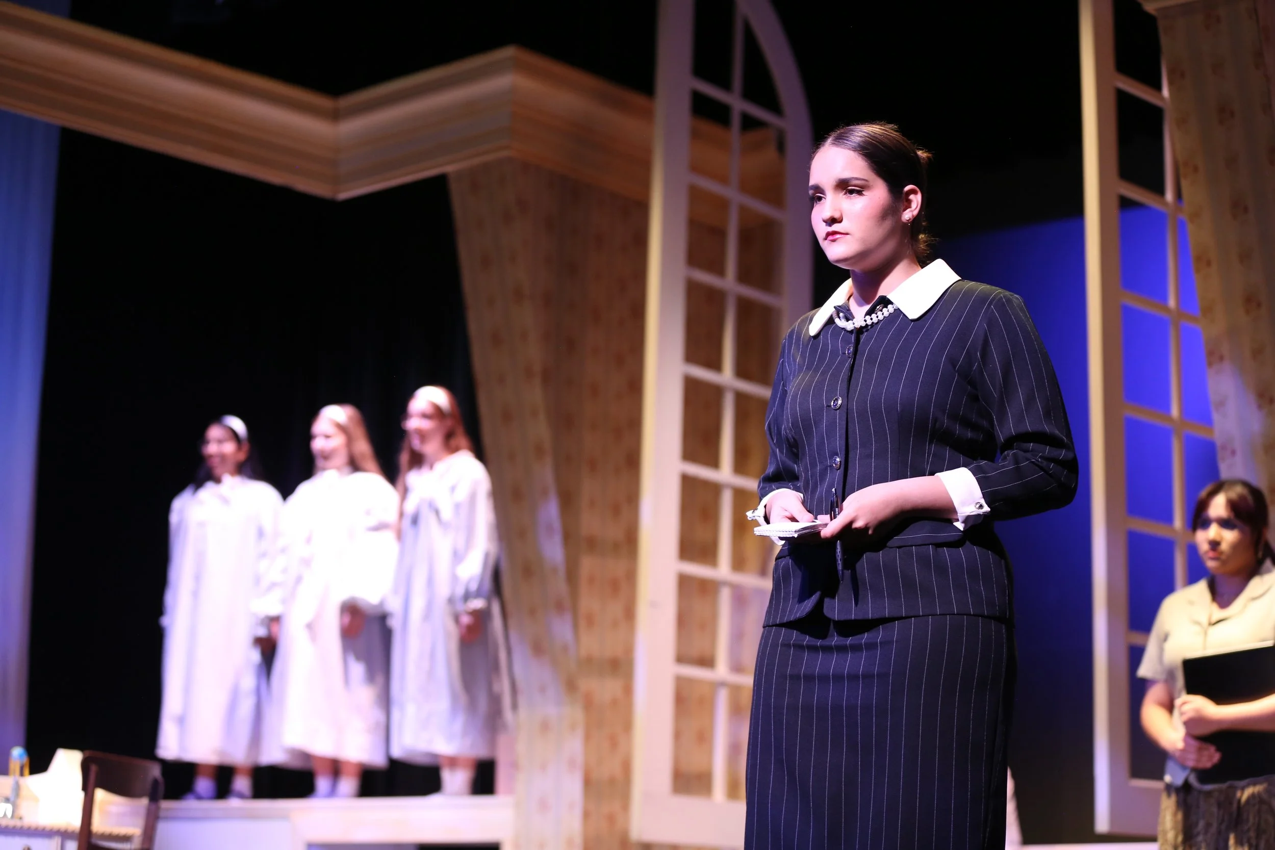 A woman in a dark pinstripe suit with a white collar and pearl necklace standing on stage, with women in white dresses and a woman in a yellow jacket visible in the background, under stage lighting with a wooden decorative set.