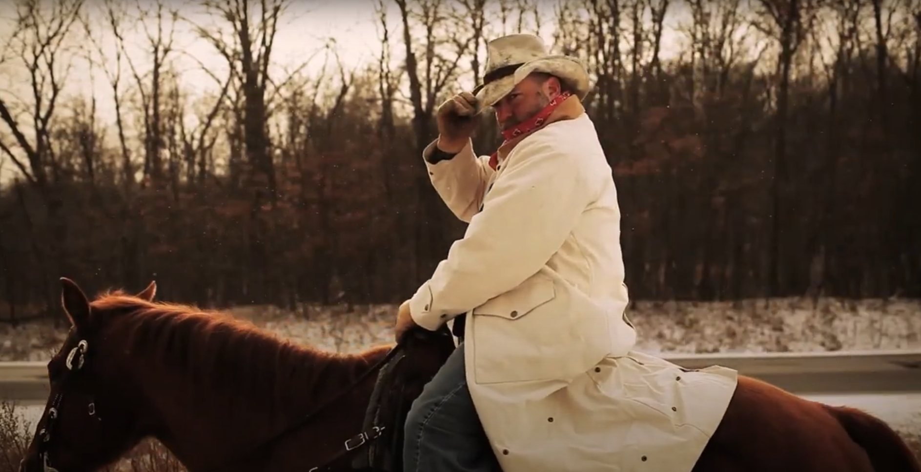 A man riding a horse on a road with a wooded background, wearing a cowboy hat, bandana, and light-colored coat during sunset.