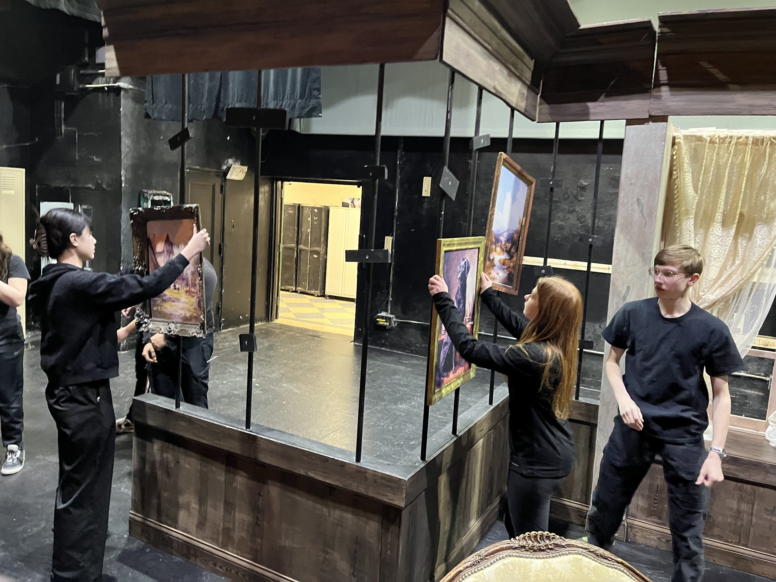Students assembling the set of a one-act play. There are low wood panel walls with pipes holding up crown molding and vintage parlor room picture hung on the pipes.