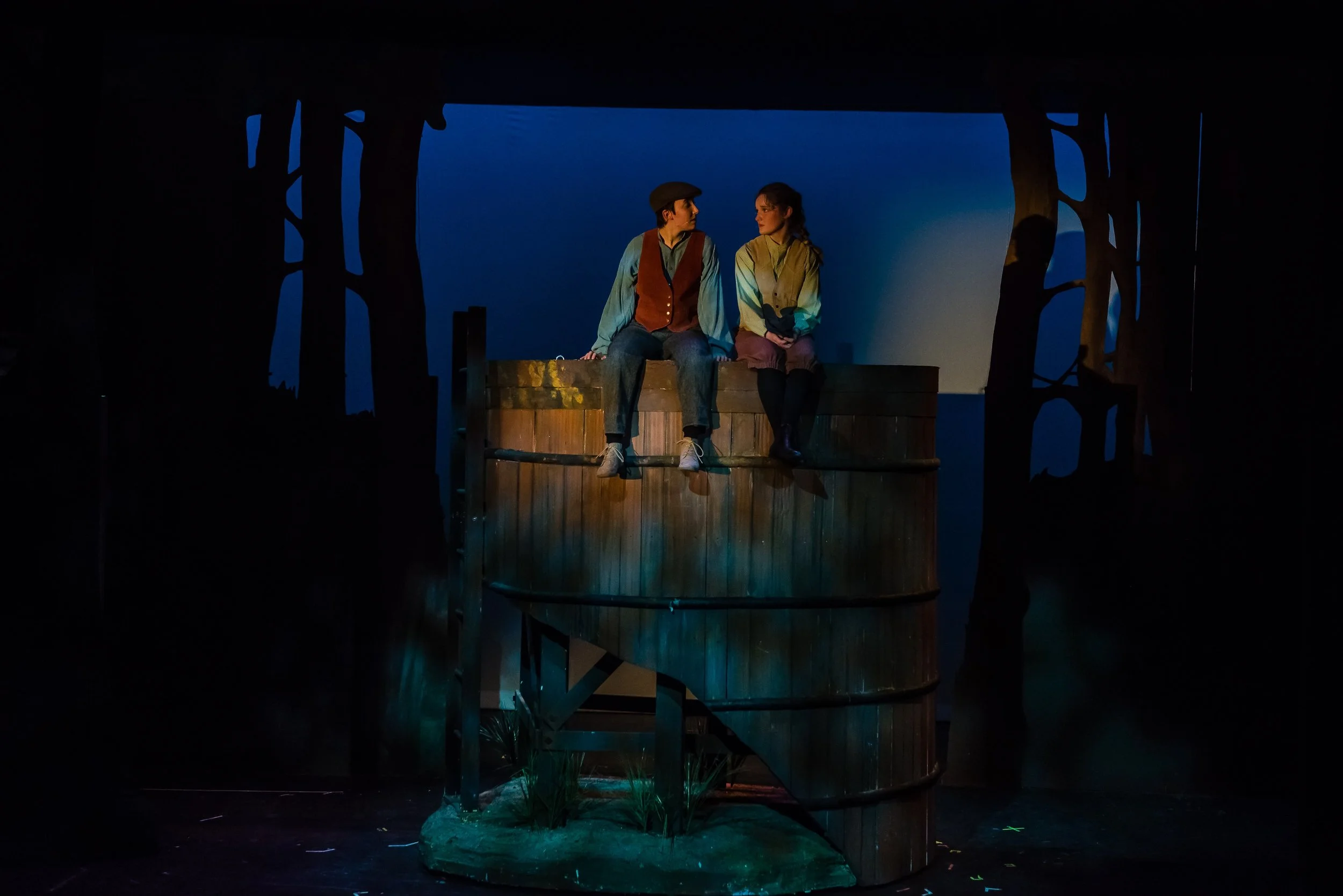 Two people sitting on a wooden platform, engaged in conversation, surrounded by dark scenery with a blue background and illuminated by stage lighting.