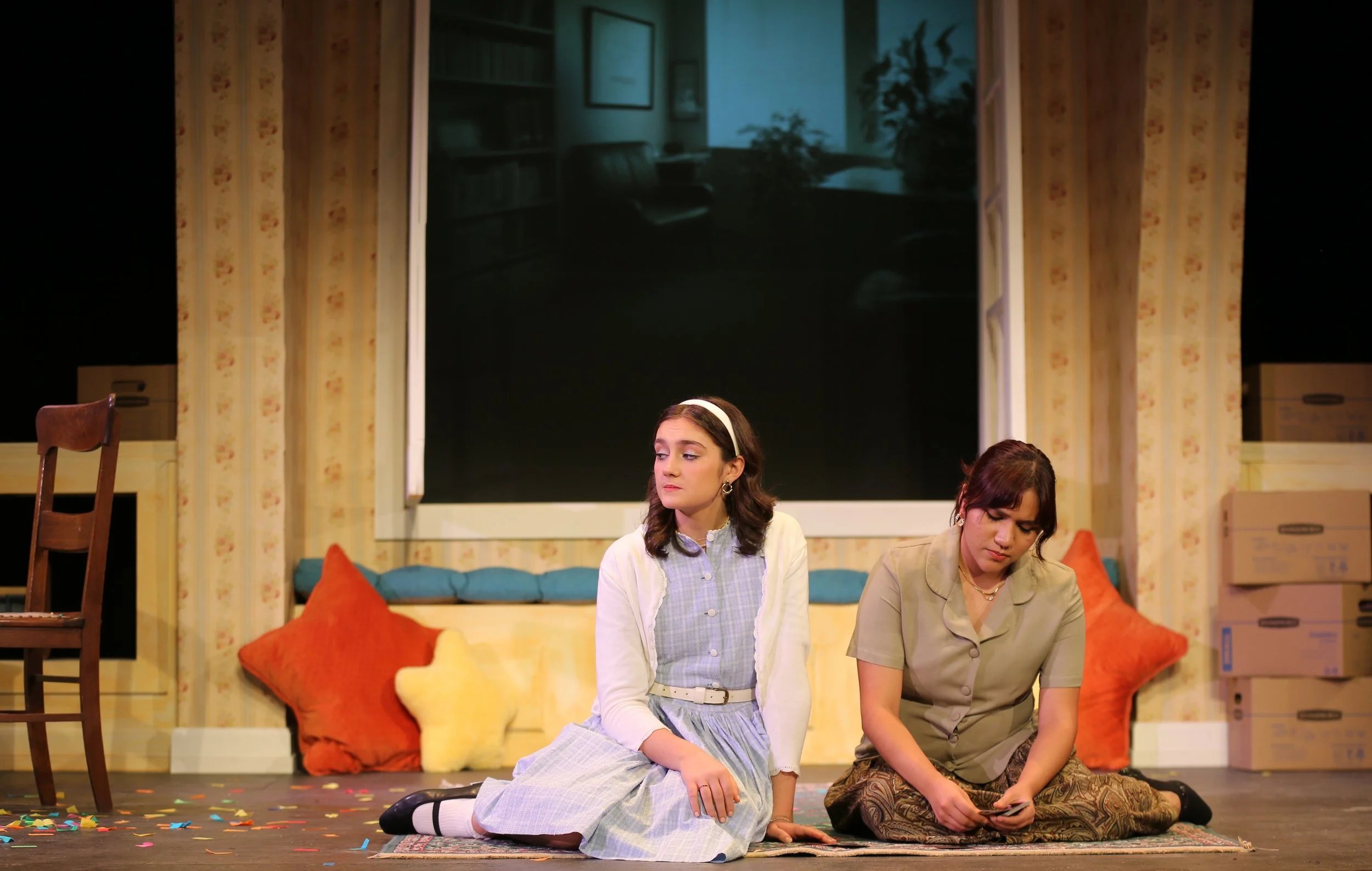 Two young women sitting on a stage in front of a backdrop of a living room set, with pillows, boxes, and a window, one scrolling on a phone and the other looking away, surrounded by colorful confetti on the floor.
