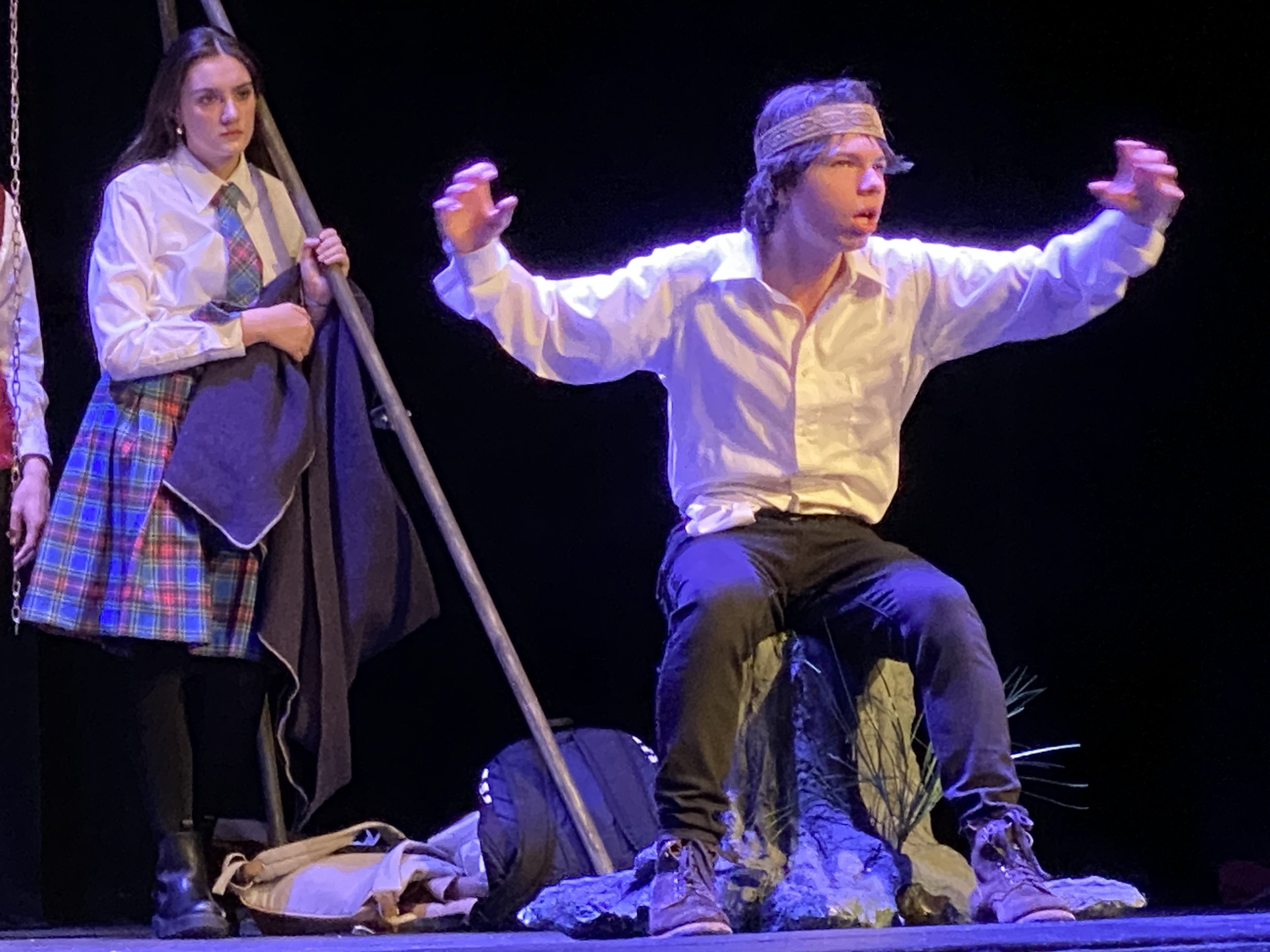 Two young actors performing on stage, one standing and holding a stick, the other sitting with arms outstretched, both dressed in costumes, with a dark background and stage props.