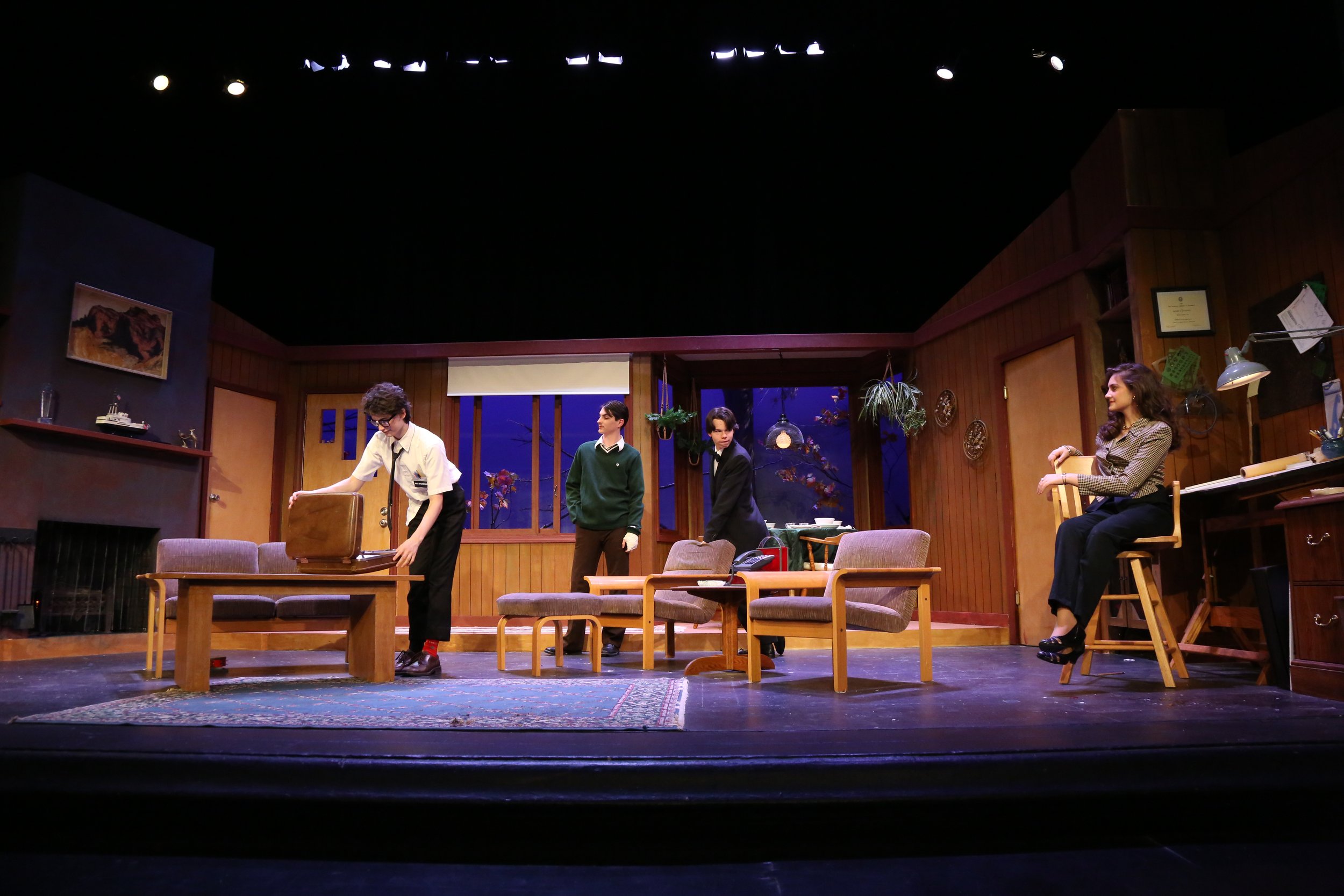Theatre stage set with four actors, some seated, others standing, with wooden furniture, a potted plant, and framed pictures on wood-paneled walls, under stage lights.
