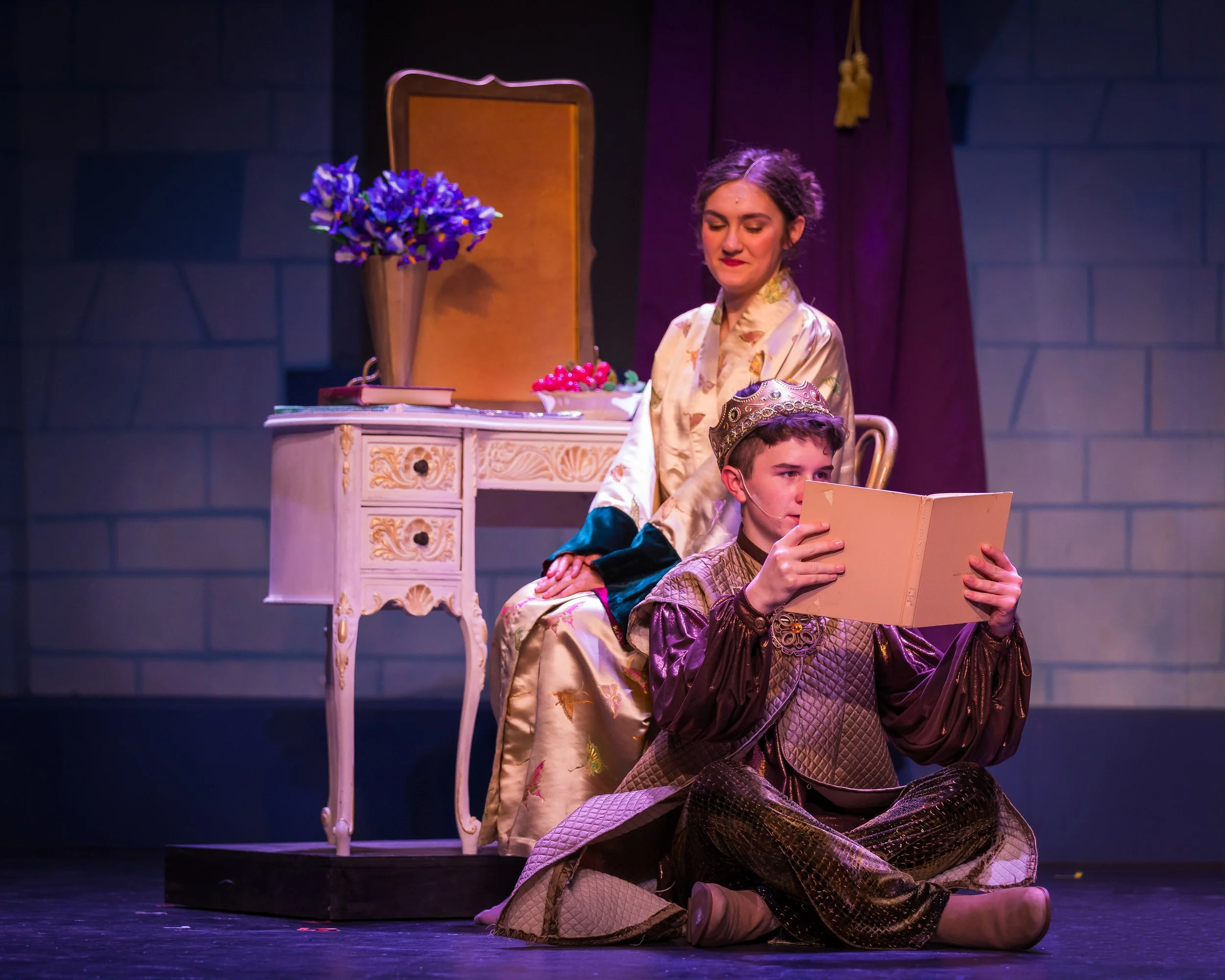 The image shows a woman with dark hair and a young boy, both dressed in ornate, patterned costumes, on a stage. The woman is sitting behind the boy, with her eyes closed and a calm expression. The boy is seated in front of her, holding a book, and ap