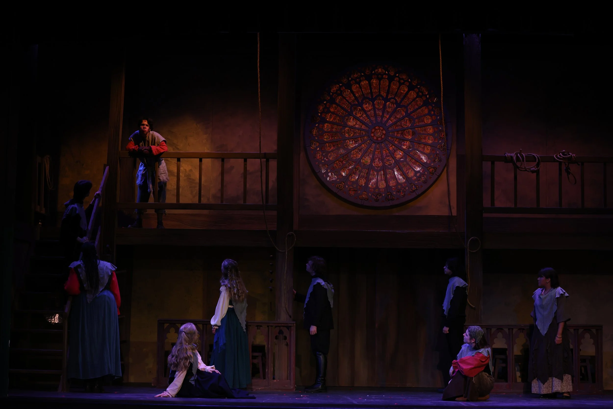 Scene from a theatrical play with actors in period costumes on stage, featuring a large circular stained glass window and ropes, with some actors on a raised platform and others on the stage floor.