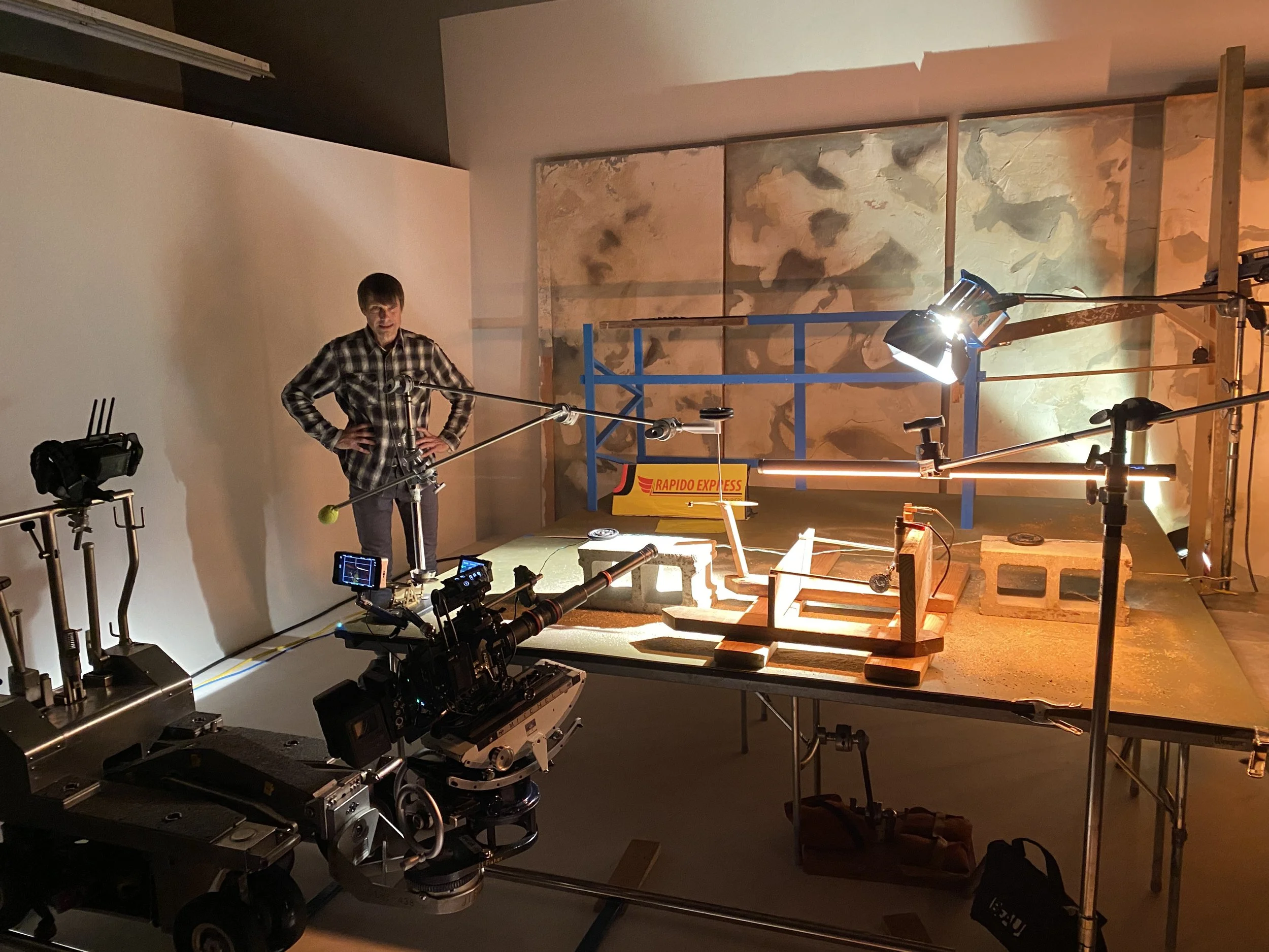 A person in a plaid shirt standing amidst filming equipment, including cameras, lights, and tracks, in a studio with a distressed wall backdrop.