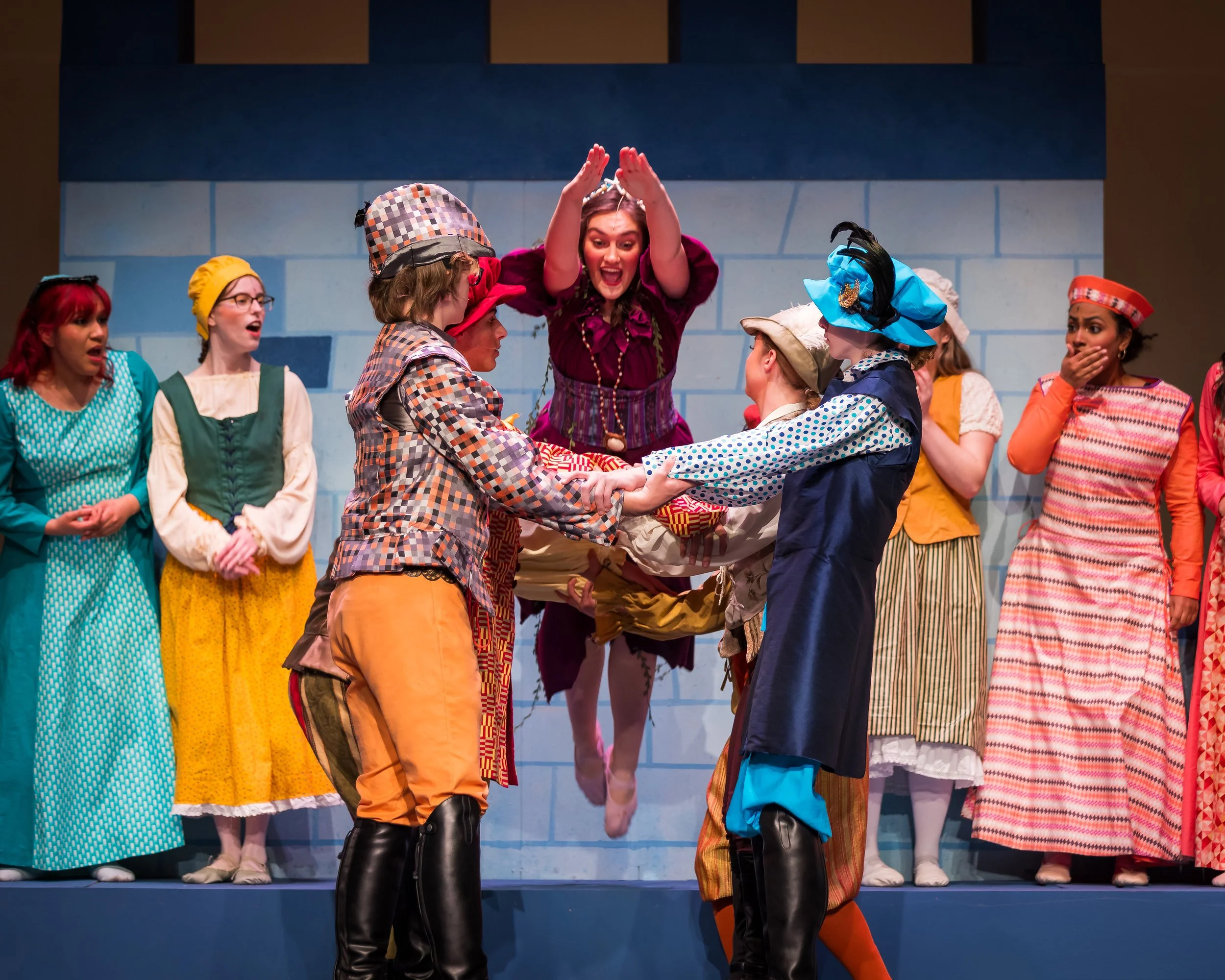 A theatrical scene featuring actors in colorful, vintage-style costumes. They are on stage with a blue brick wall backdrop. One woman is mid-air, jumping with her arms raised, while others around her hold hands, singing and acting out a scene.
