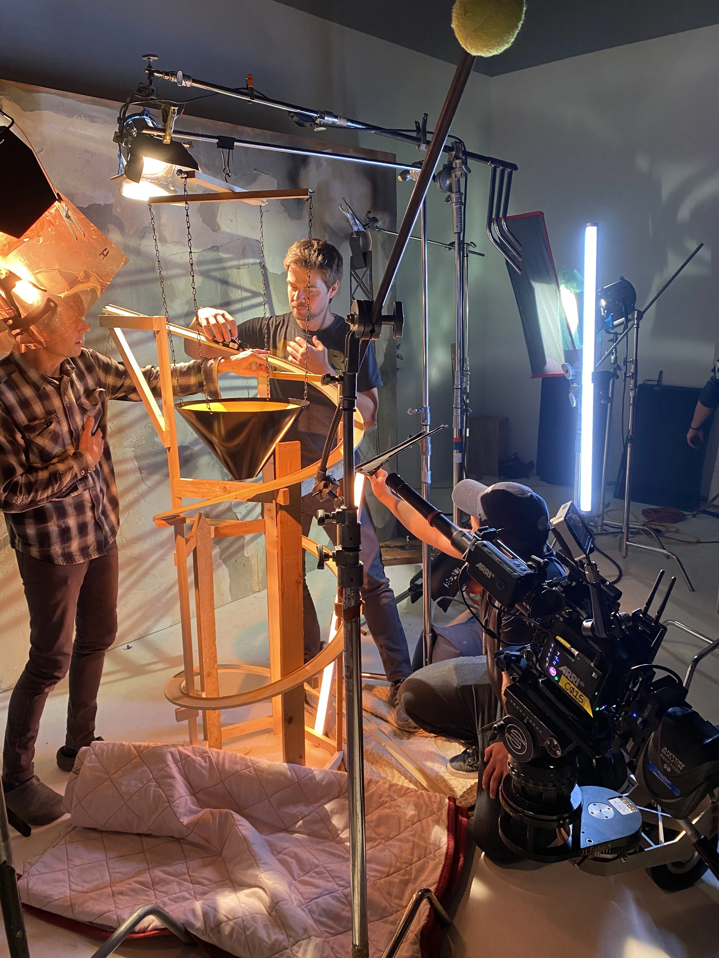 A film set with lighting equipment, a camera, and crew members preparing a scene with a wooden structure.