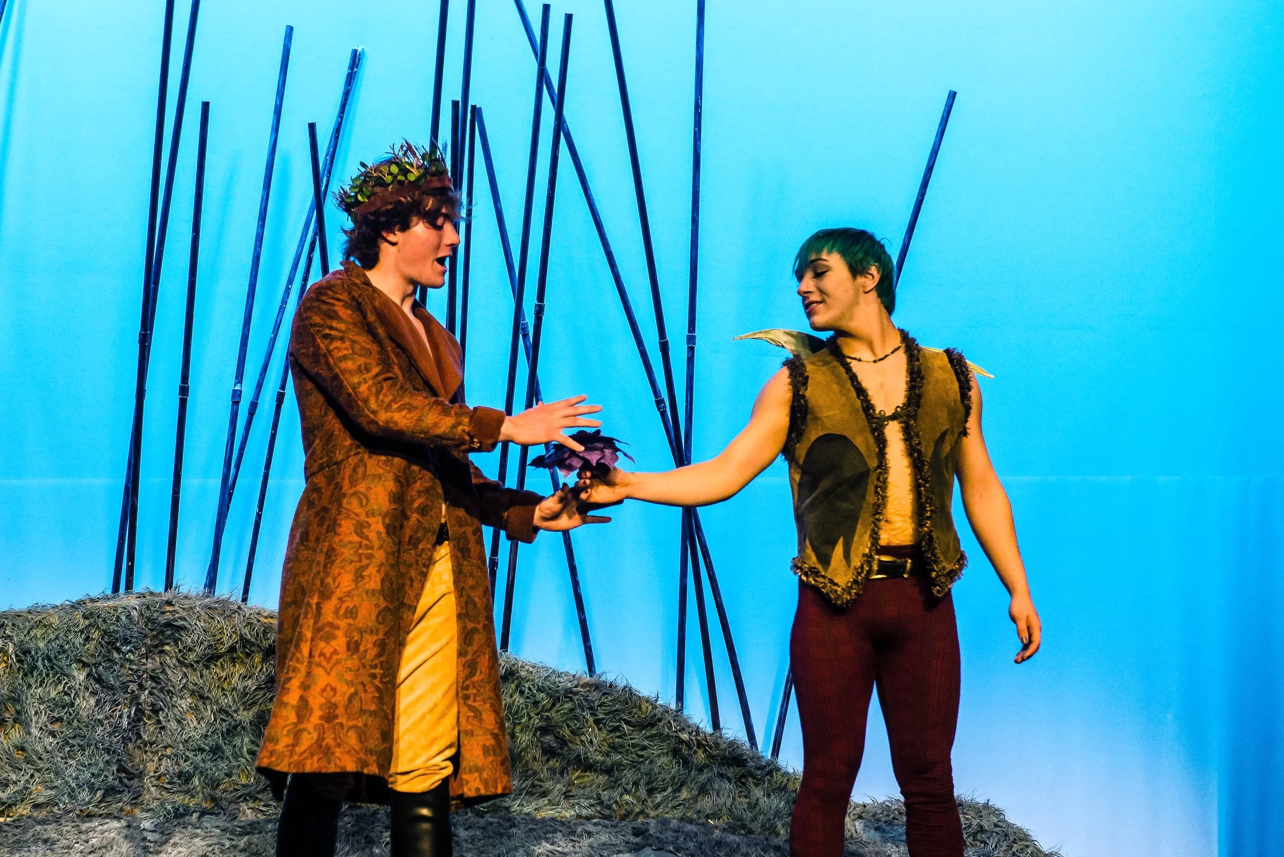 Two actors on stage dressed as characters from a fantasy or fairy tale setting, exchanging a handshake. The background is a blue sky with bamboo-style poles, and they stand on a textured surface resembling grass or moss.