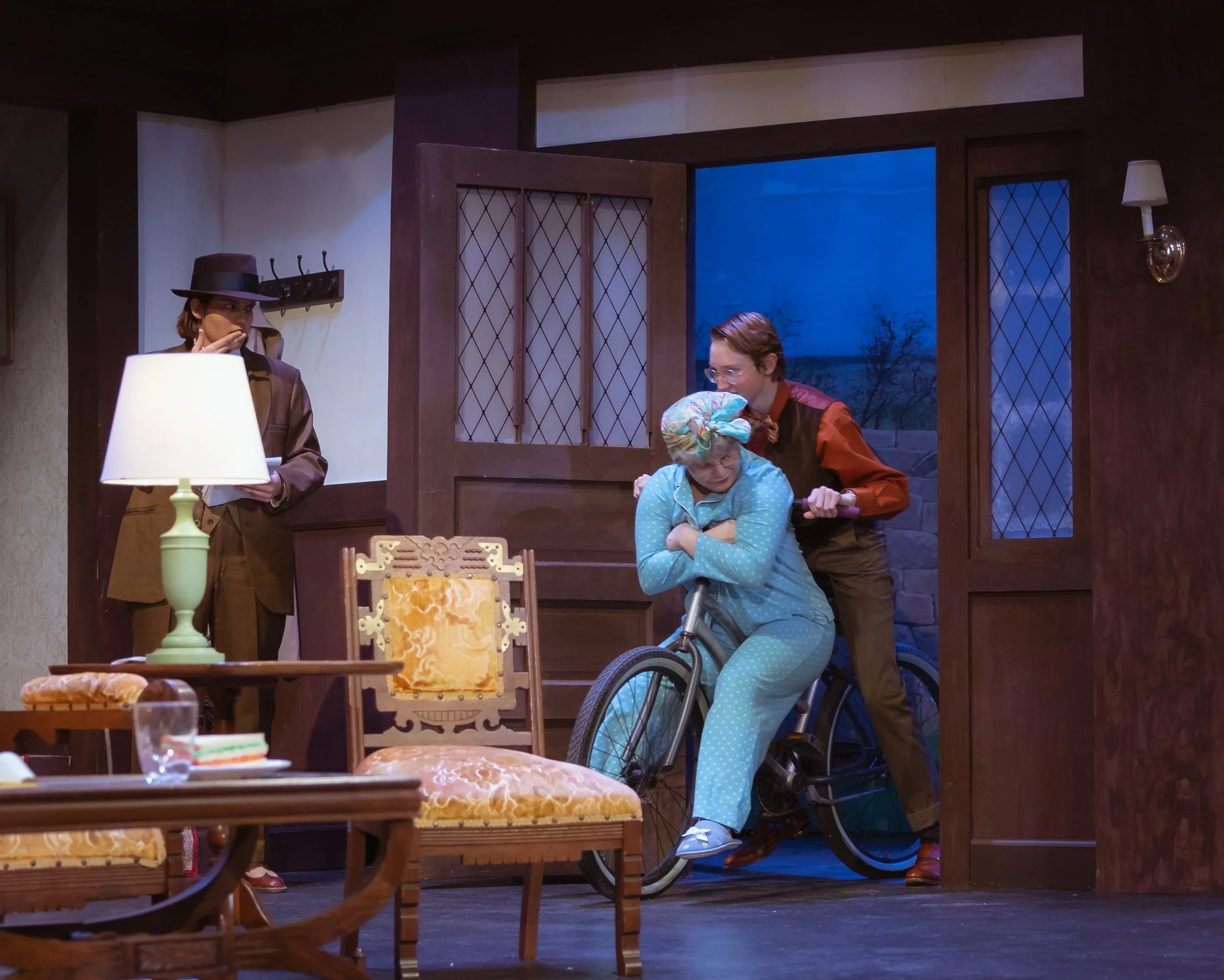 Stage scene from a play with three actors, two are on a bicycle indoors with wooden frames and a blue evening sky visible through the window.