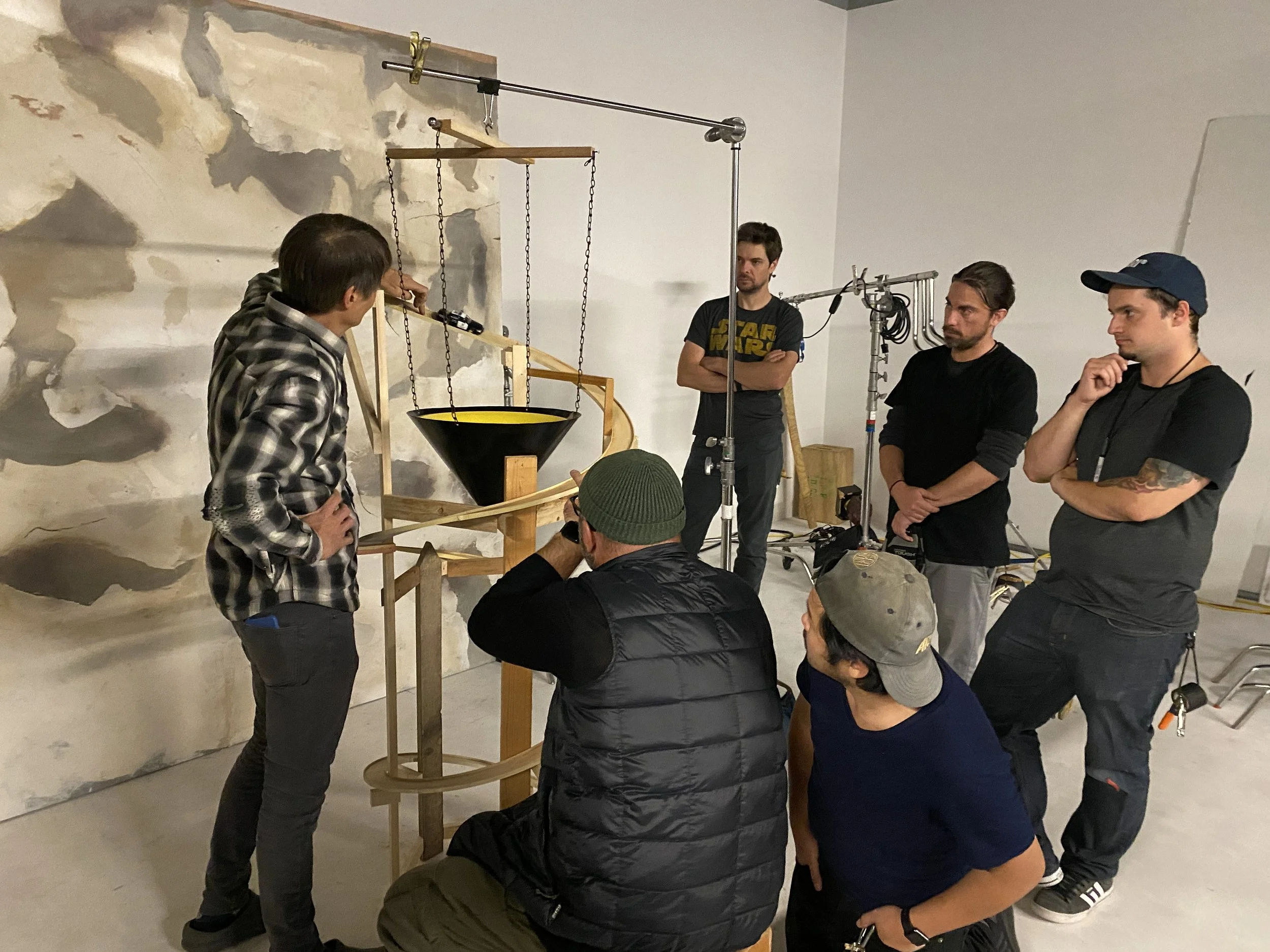 A film crew gathered around a wooden structure with chains and a funnel in an indoor space with a painted wall background.