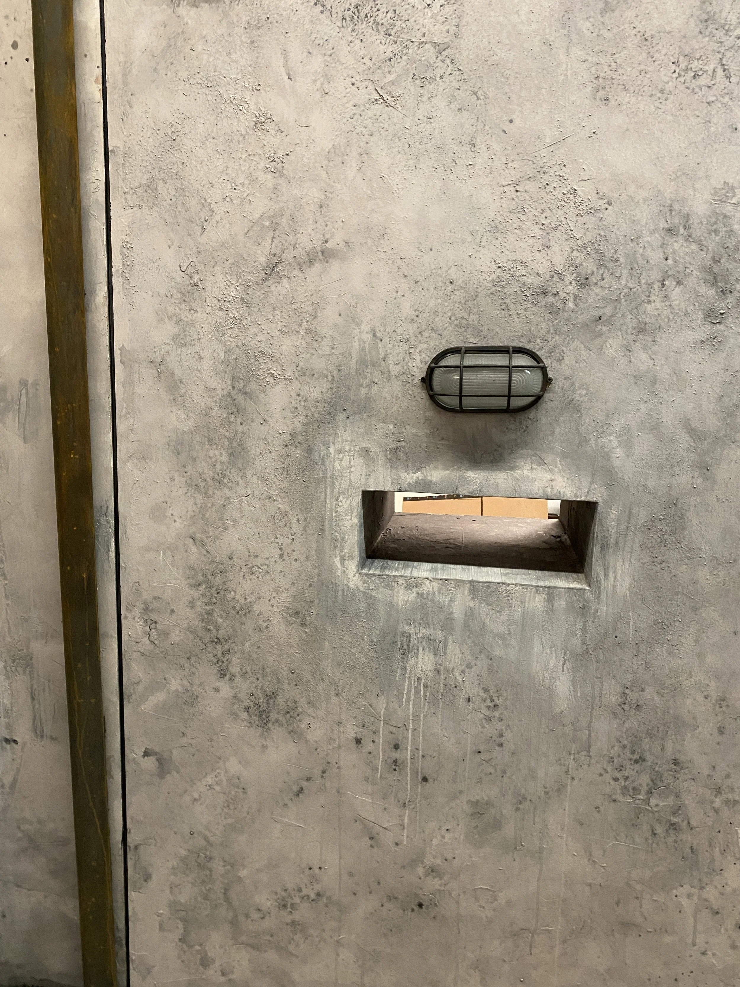A wall with a textured concrete surface, a rectangular cutout or opening, and a wall-mounted light fixture with a grid protective cover.
