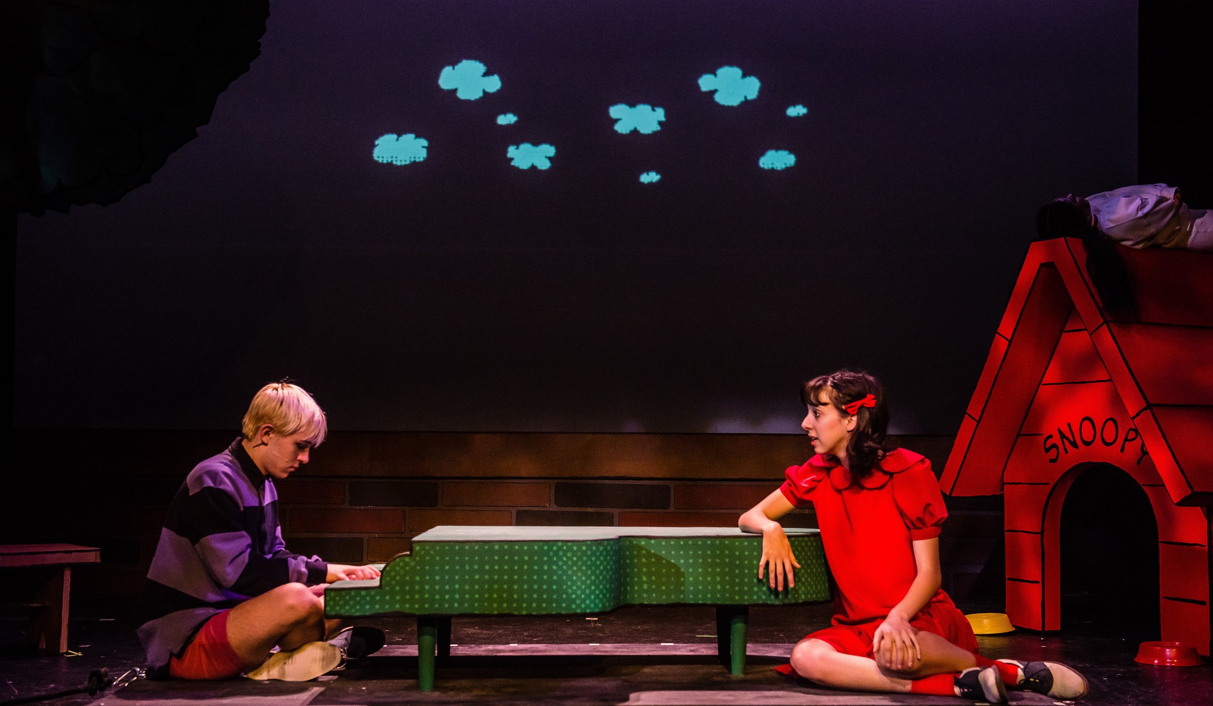 Two children performing in a play, one playing a piano and the other sitting on the floor in a red costume next to a doghouse labeled 'SNOOPY', on a stage with a dark background and a cloud projection.