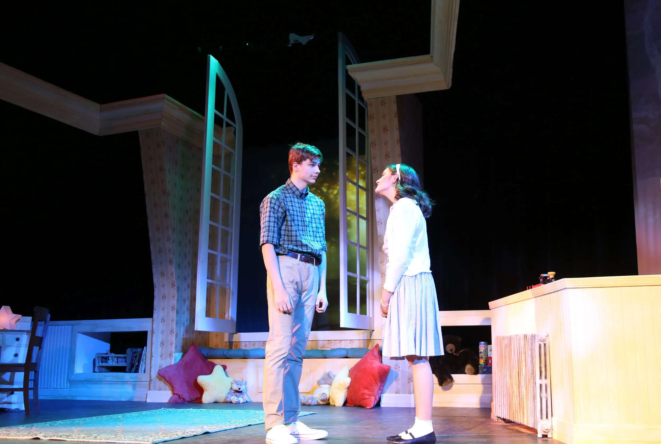 Two young actors, a boy and a girl, stand face-to-face on a stage set resembling a cozy bedroom, with large windows, bed, pillows, and stuffed animals, during a theatrical performance.