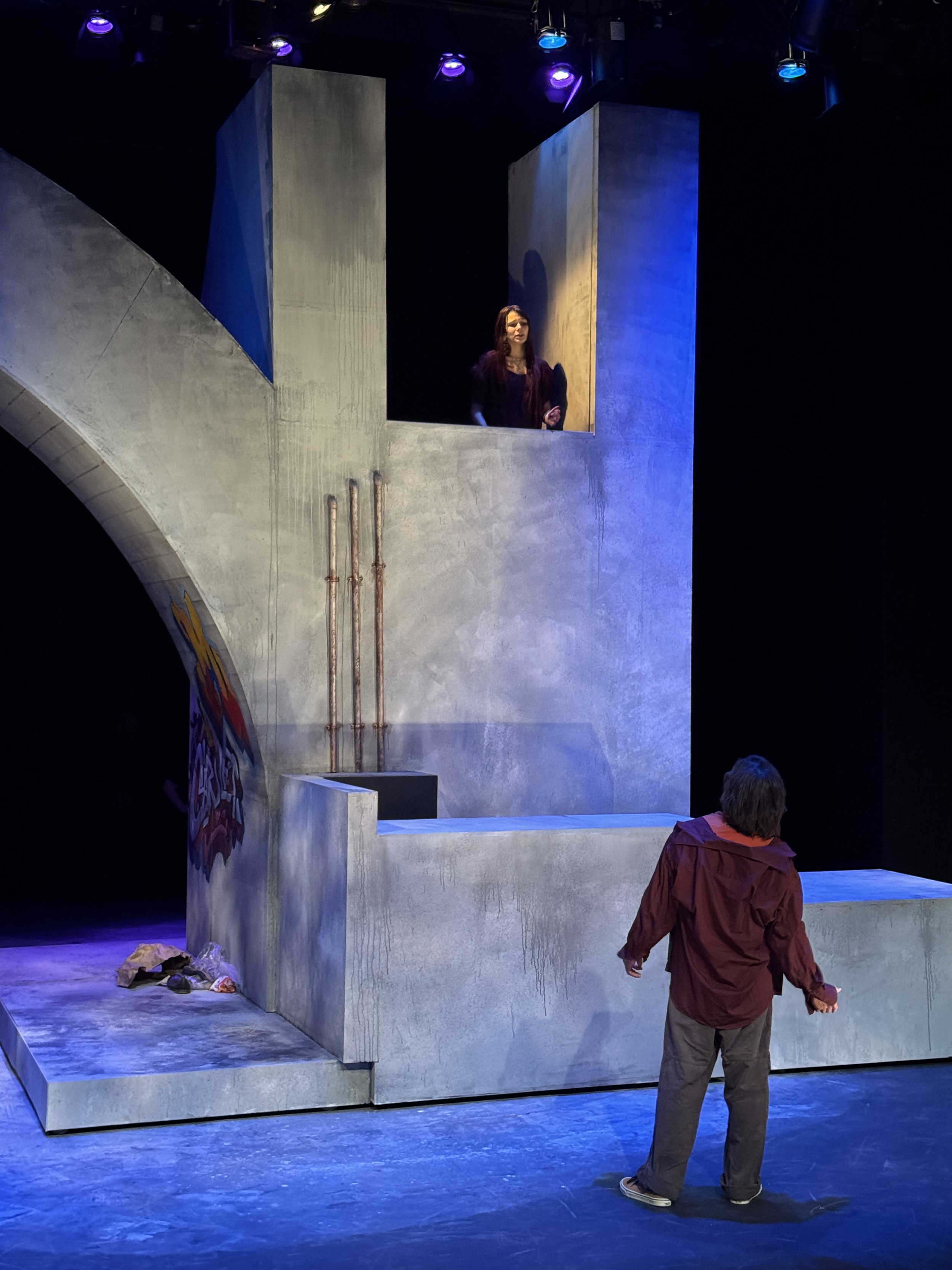 A woman with long dark hair standing inside a concrete structure on a stage, looking forward. The structure resembles part of a bridge with vertical supports and an arch. Two people are visible in the foreground, one wearing a red jacket with gray pa