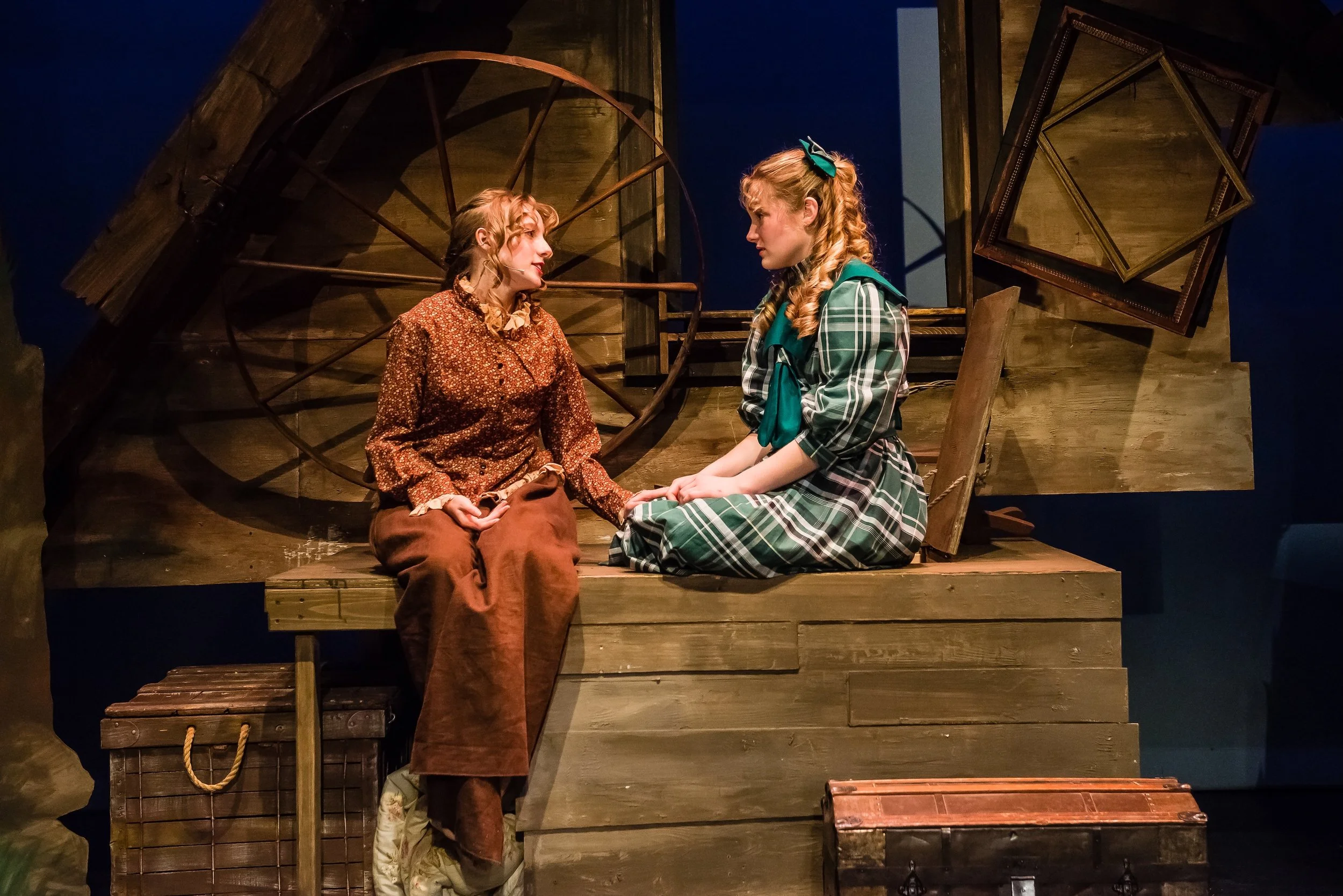 Two women with blond curly hair sitting on a wooden platform, facing each other, holding hands. One woman wears a patterned blouse and brown pants, the other wears a plaid dress with a teal scarf and bow. The background shows a rustic wooden structur