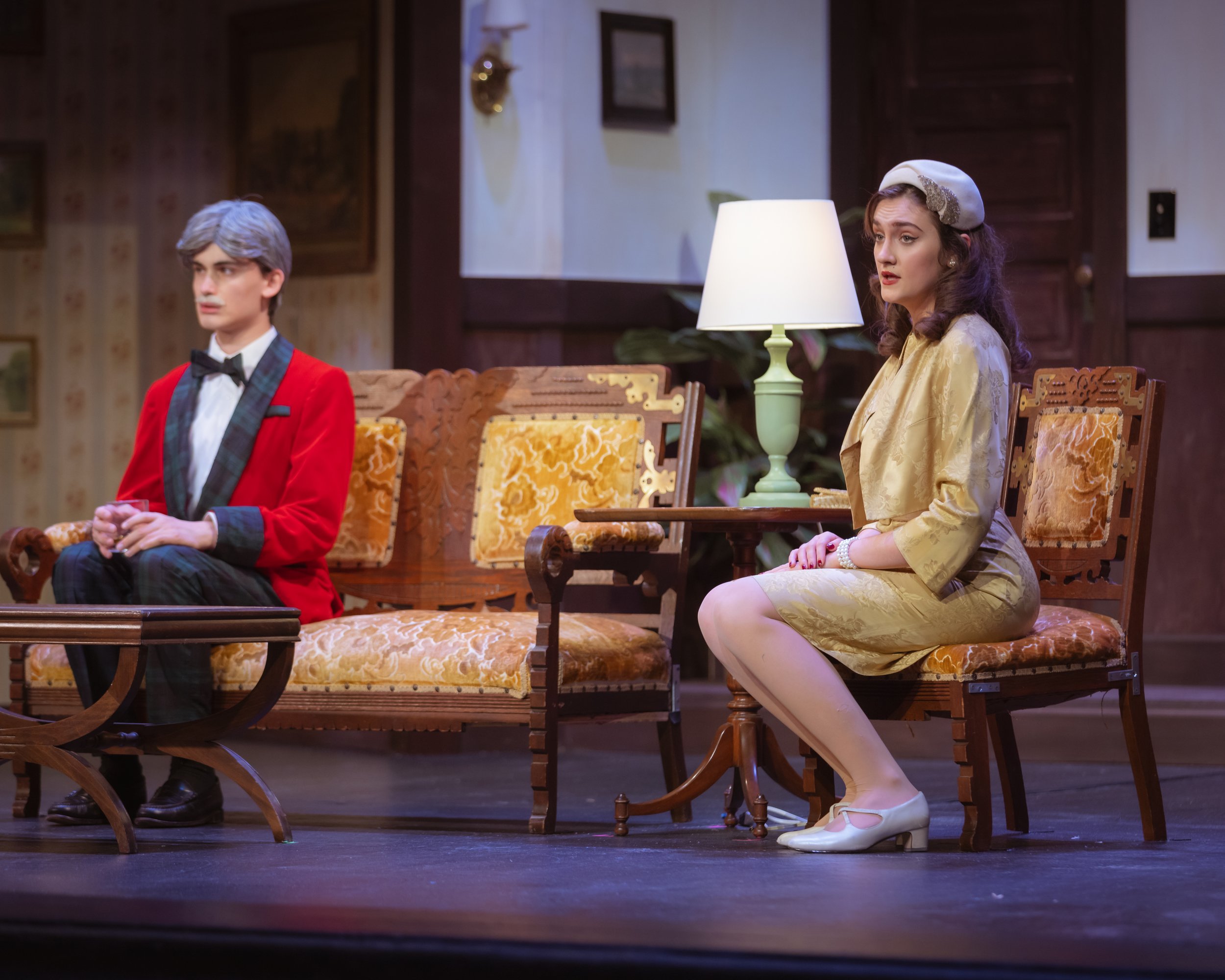Two actors, a man with gray hair and a woman with long brown hair, sitting on ornate wooden chairs in a living room setting. The man is wearing a red jacket and black bow tie, while the woman is wearing a yellow dress, white stockings, and a white ha