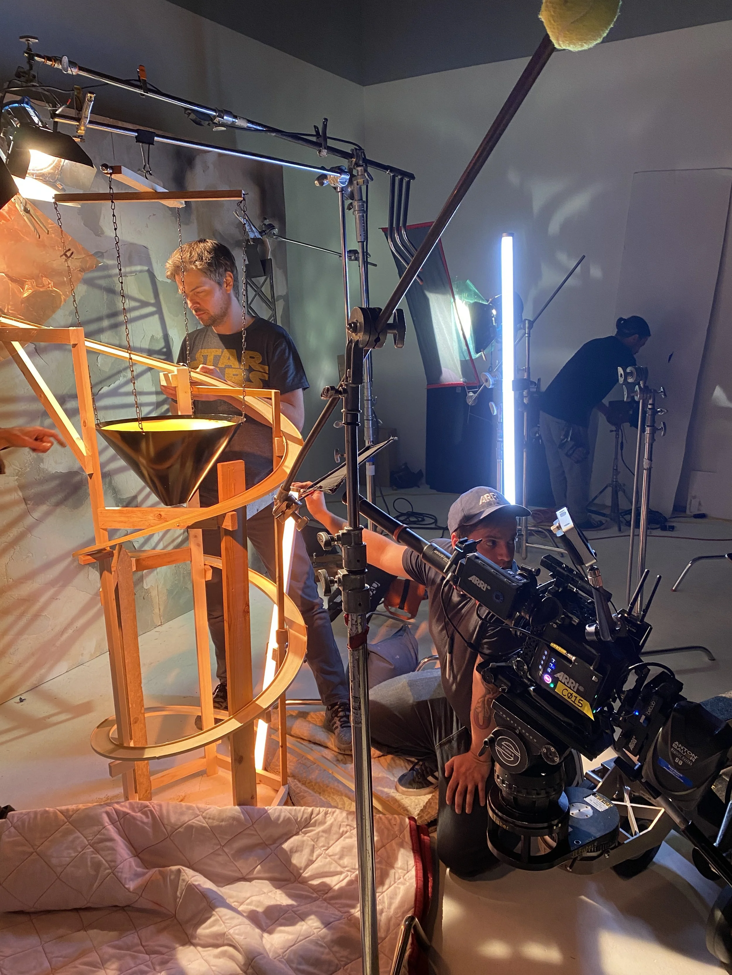 Film crew setting up a shot with a camera, lighting, and a wooden roller coaster model with a yellow funnel at the top, inside a room with studio equipment and crew members.